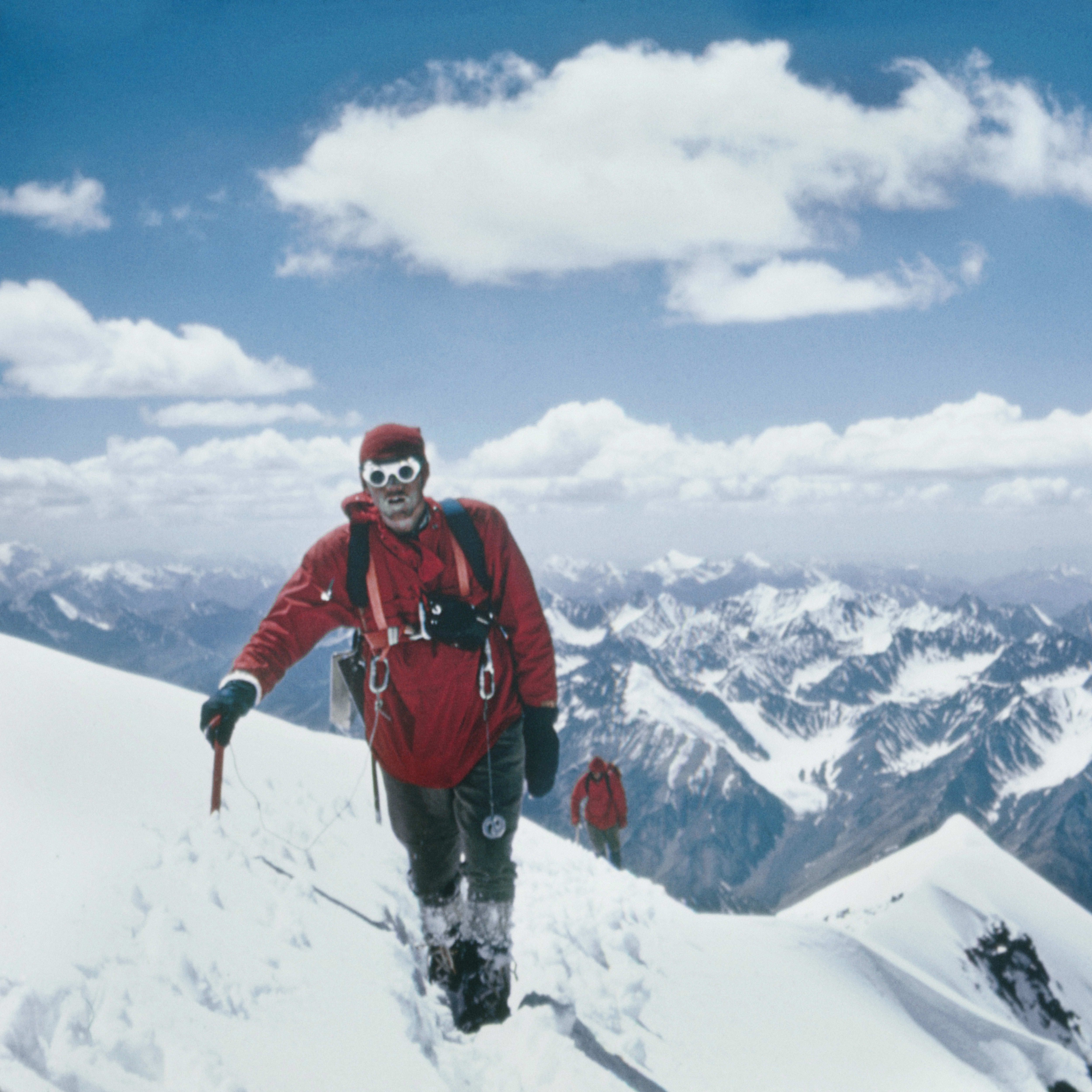 A man in red clothing, wearing goggles, is hiking across a snowy mountaintop. The mountains in the background are covered in snow. The sky is blue with white clouds.
