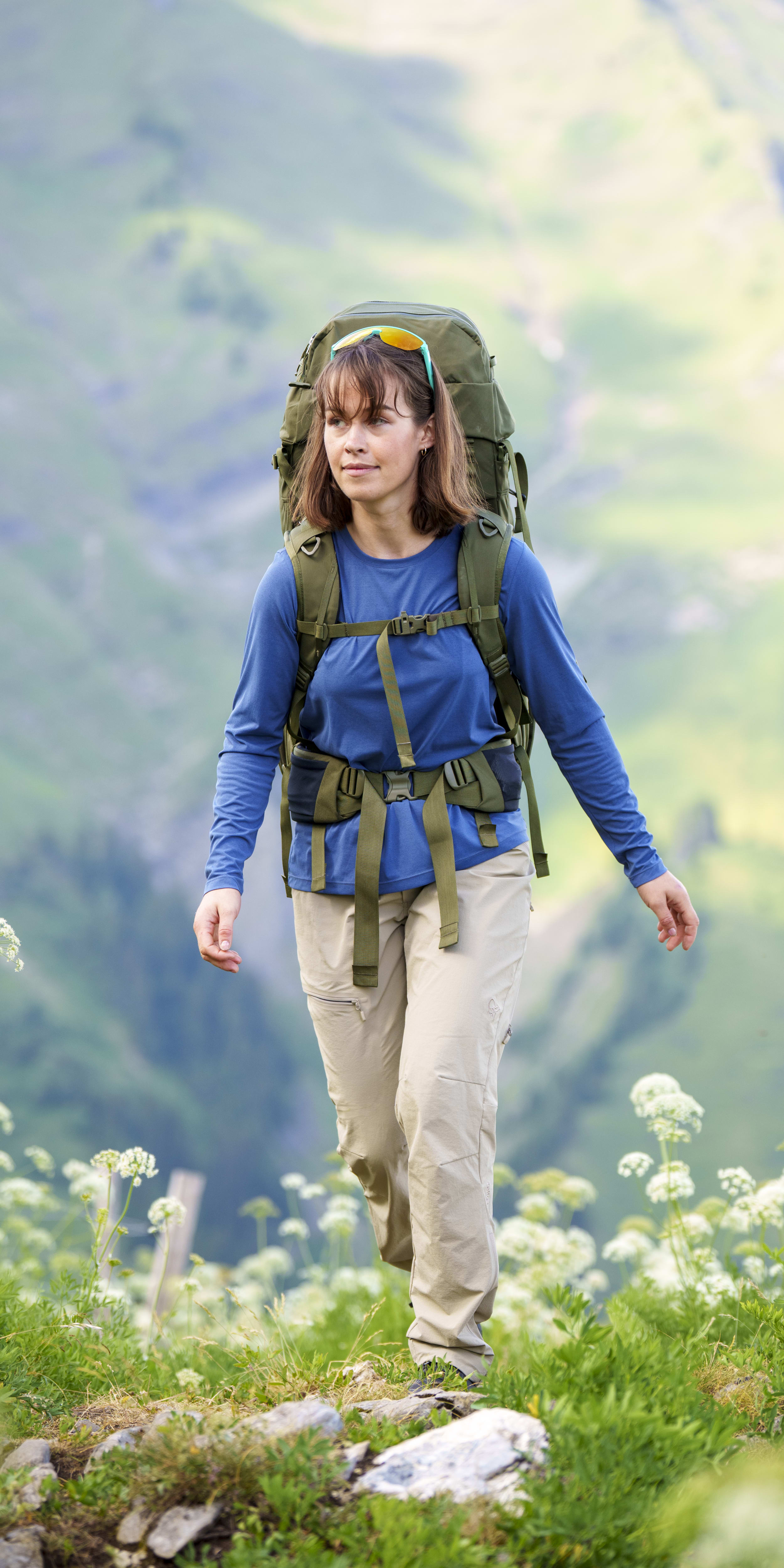 A woman hikes downhill with a large backpack. She wears a blue long sleeve shirt and beige Norrøna pants, sunglasses on her head, with mountain scenery in the background.