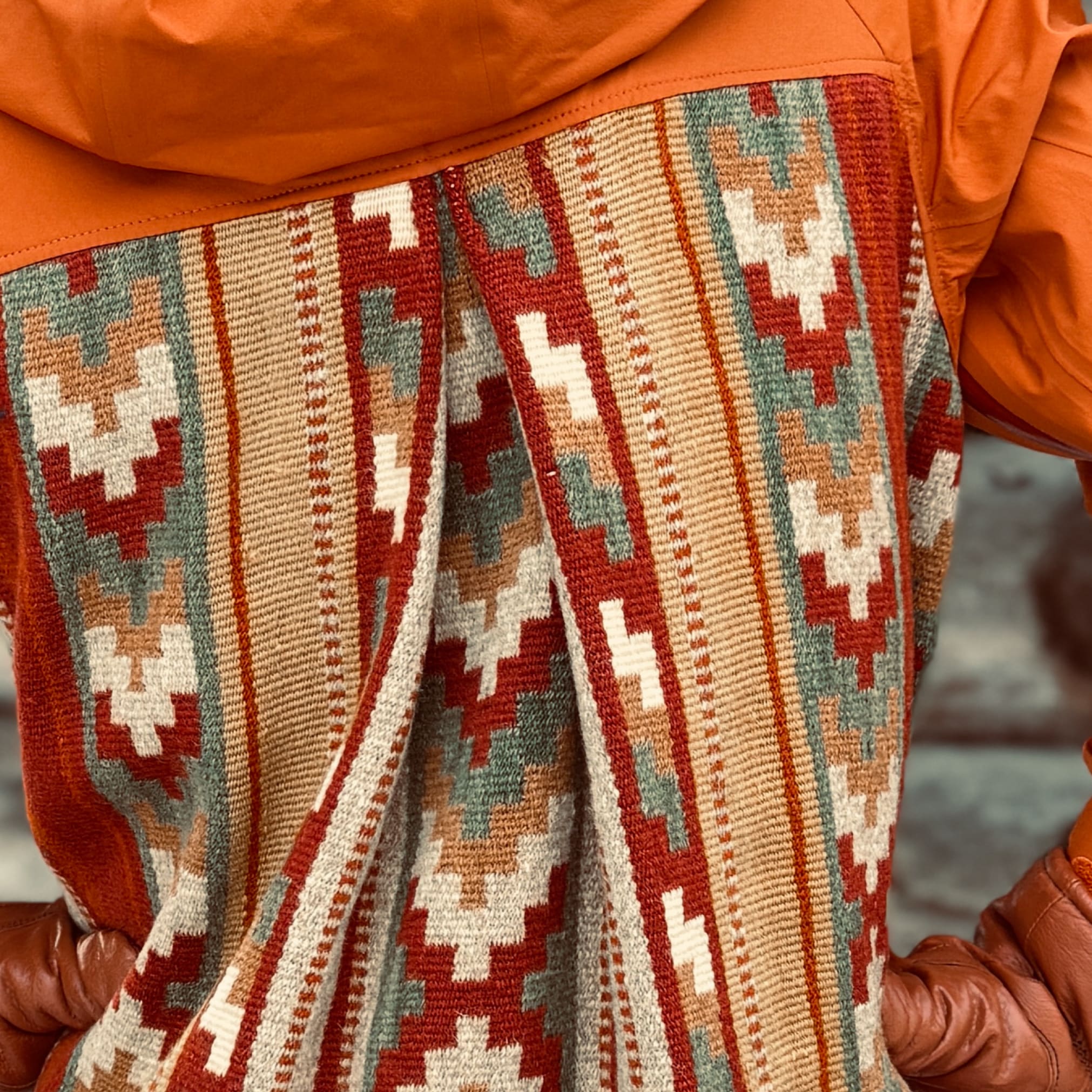 Back view of person in orange Norrøna jacket with southwestern patterned textile panel, wearing brown leather gloves, against a wooden backdrop.