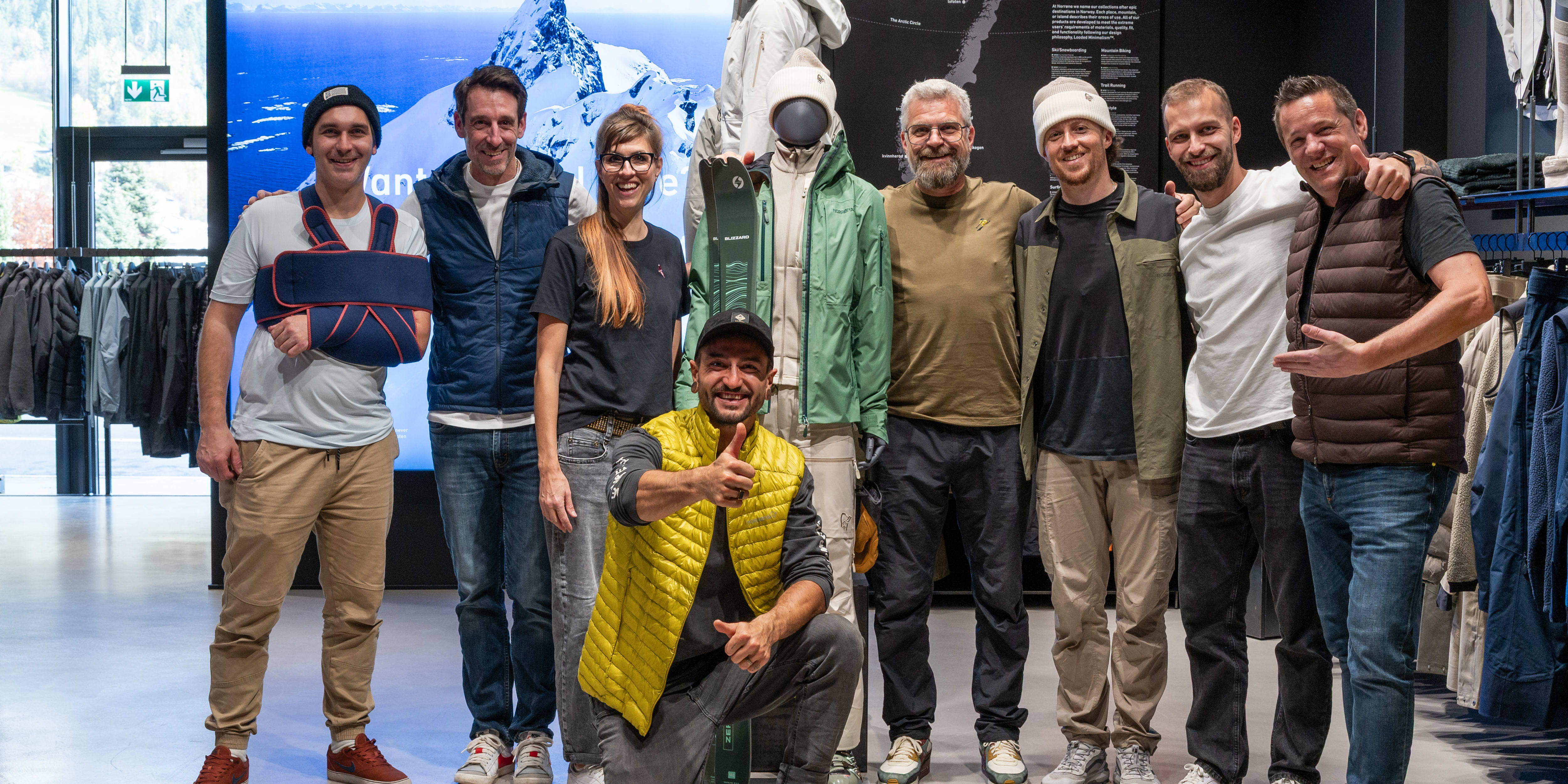 Group of 8 smiling people posing indoors with Norrøna clothing on mannequins and on some people in front of a mountain image.
