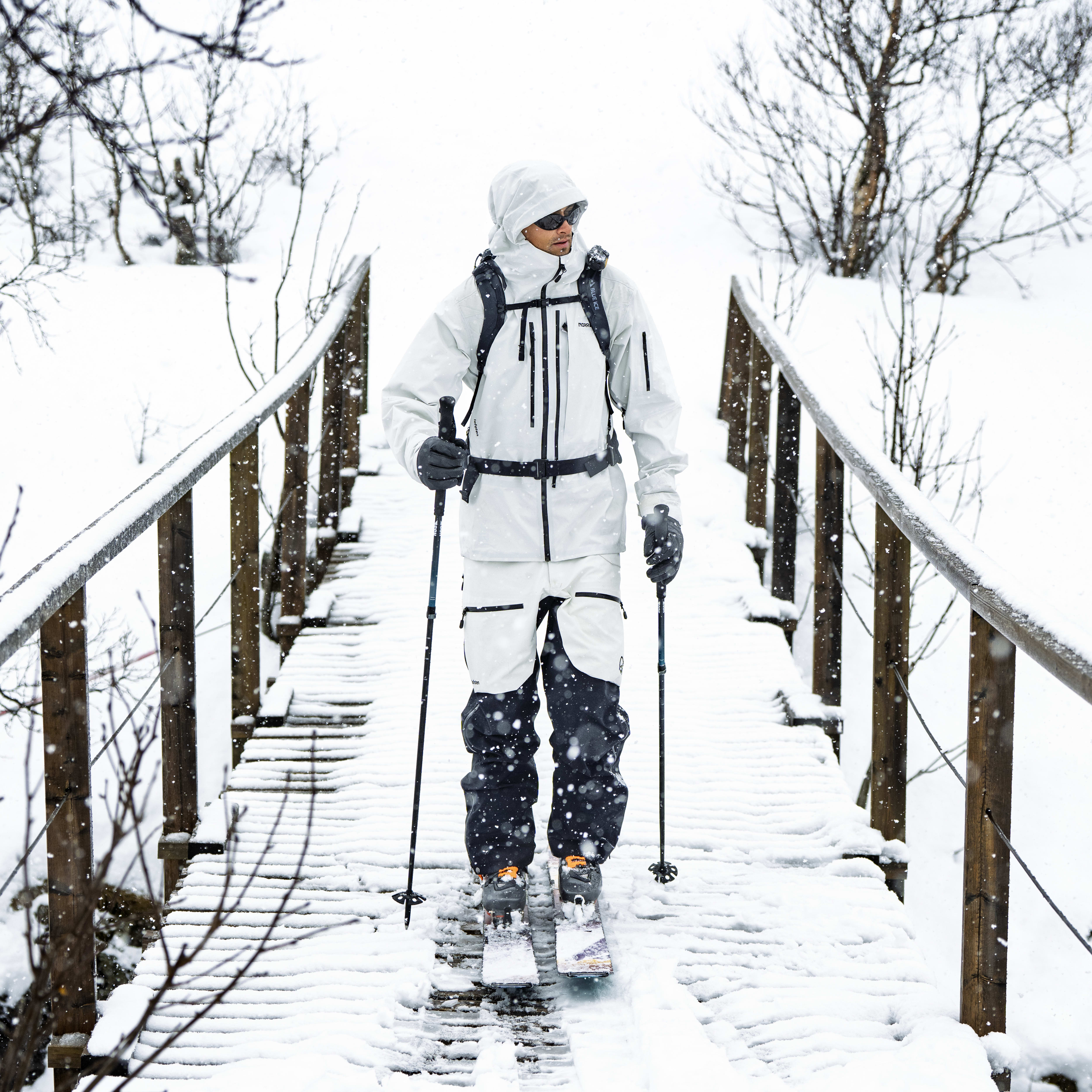A person in a white Norrøna ski suit walks on a snow-covered wooden bridge.