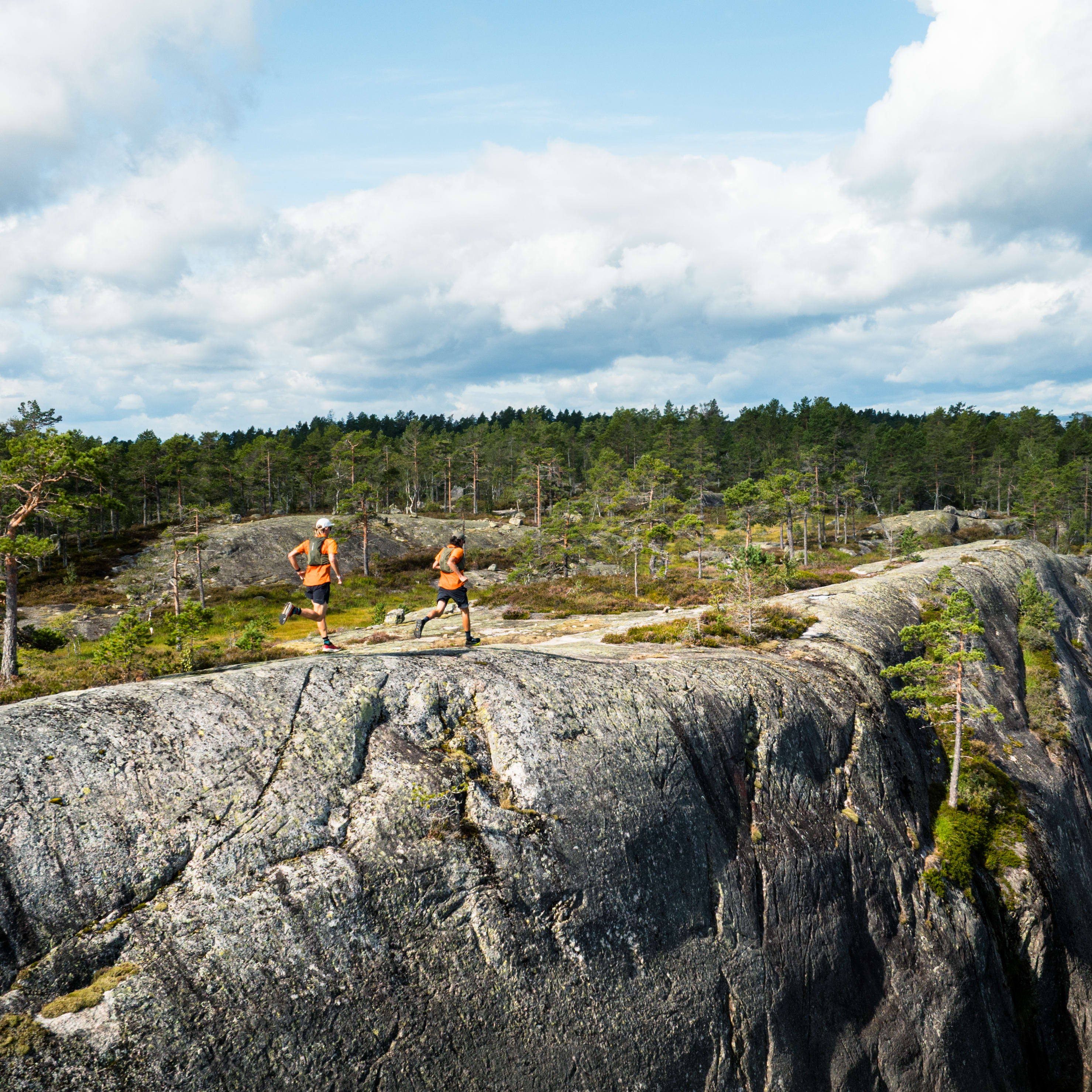 Two runners, wearing Norrøna shirts, run on a rocky ridge overlooking trees. Cloudy blue sky above.