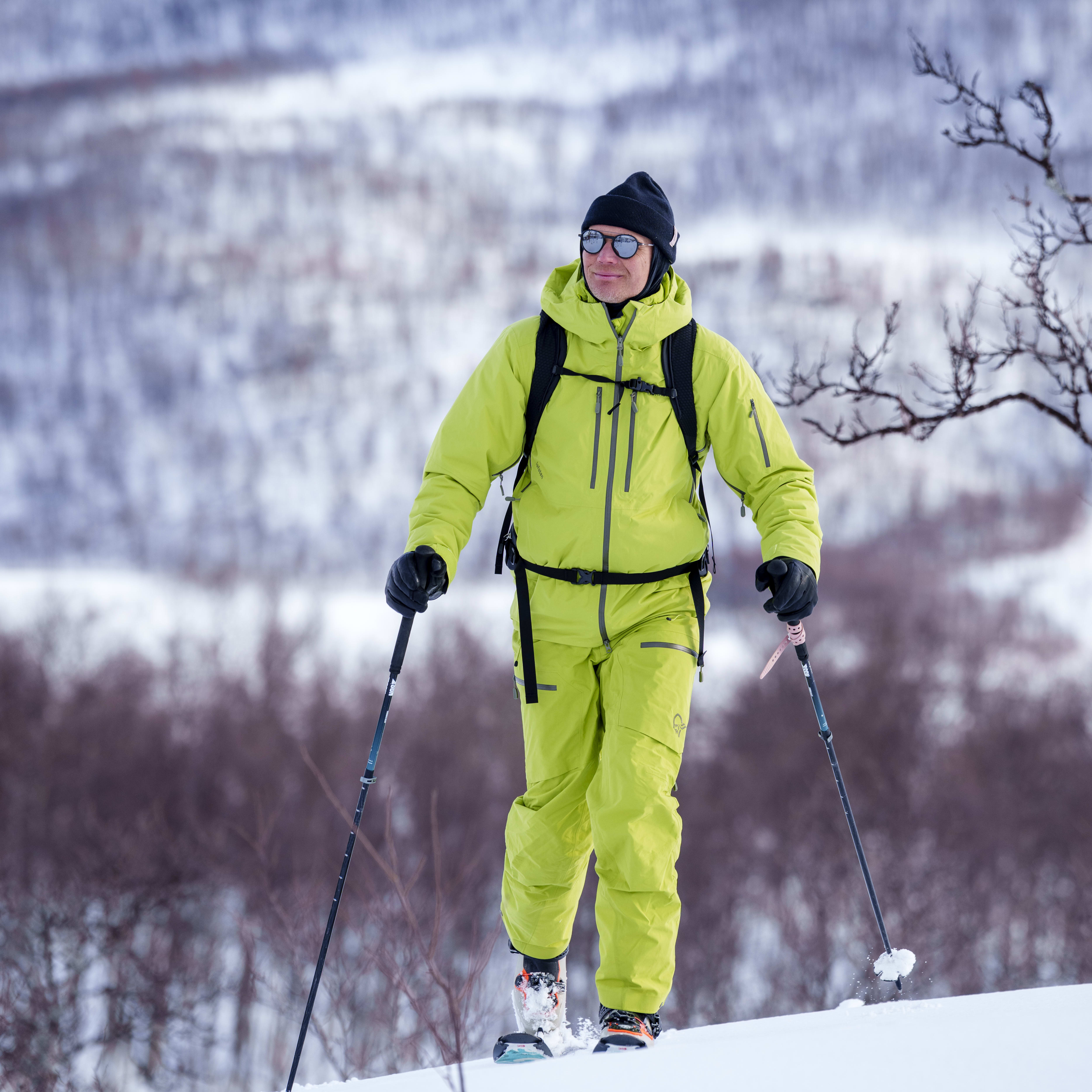 A man in a lime green Norrøna ski suit and beanie skis uphill on a snowy slope with trees in the background.