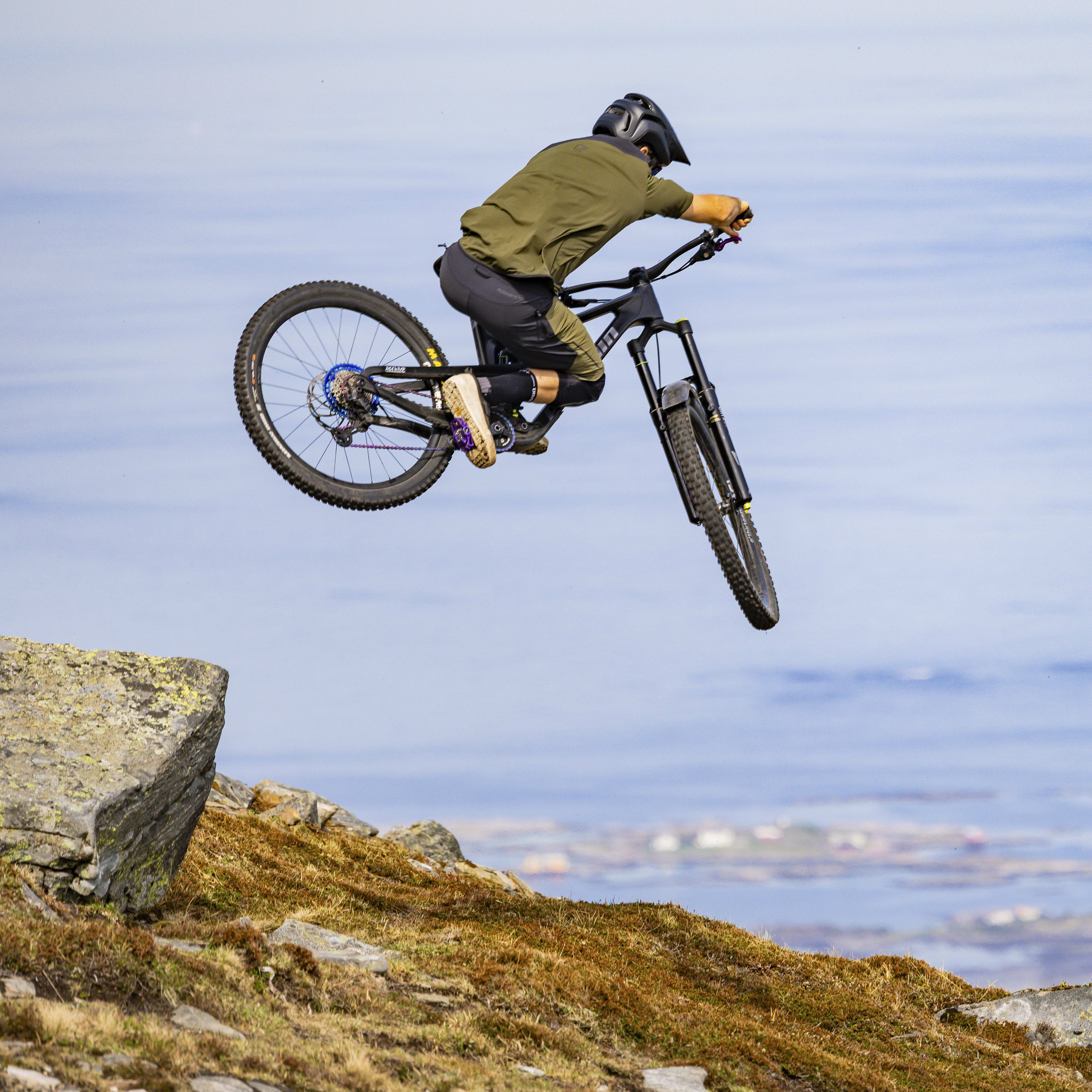 A person in Norrøna clothing performs a mountain bike jump.  The bike is in mid-air, with the rider's legs extended.  The background shows a coastal landscape with a boat in the distance. 
