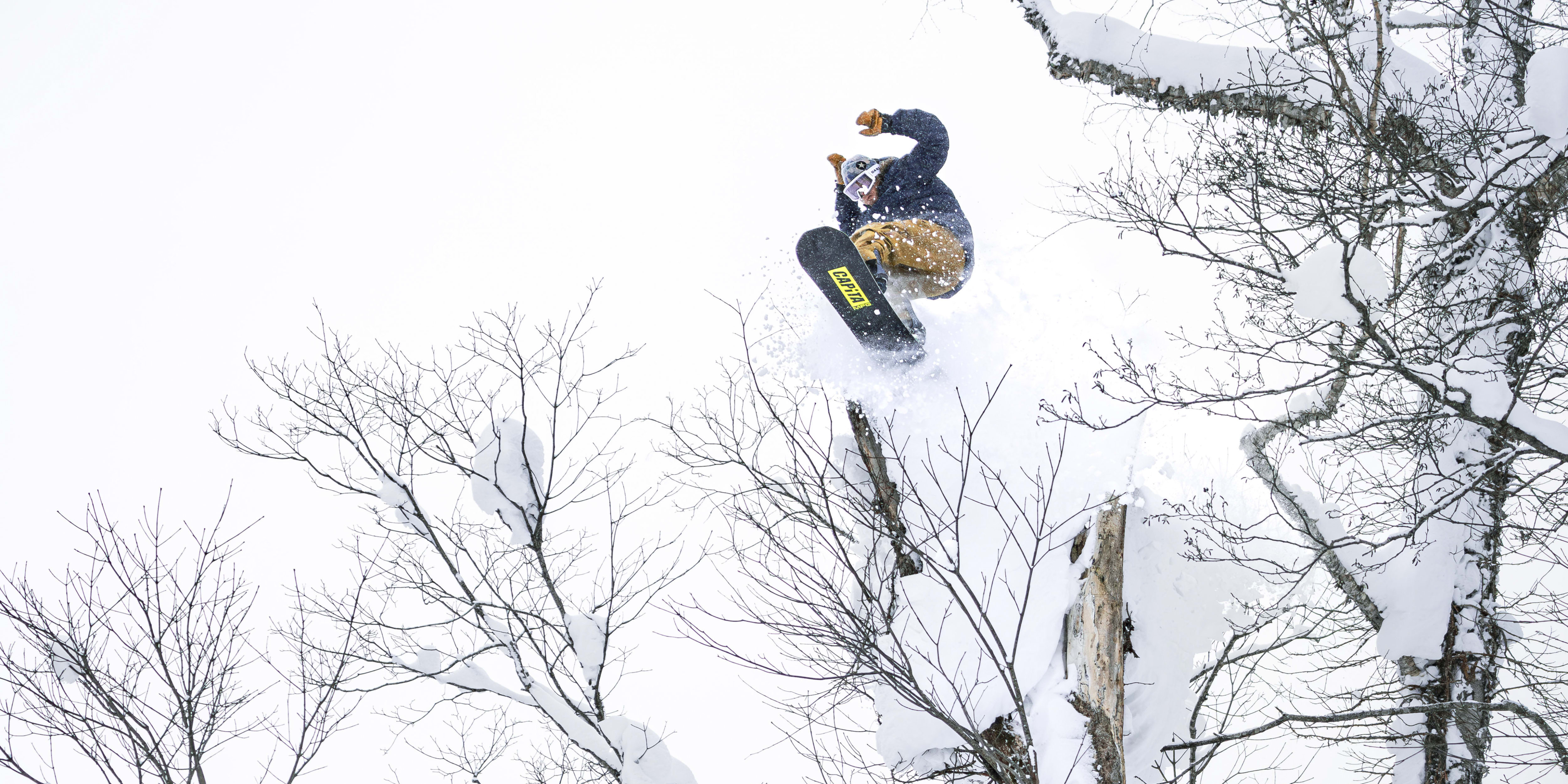 A snowboarder jumps in the air over snow covered trees.