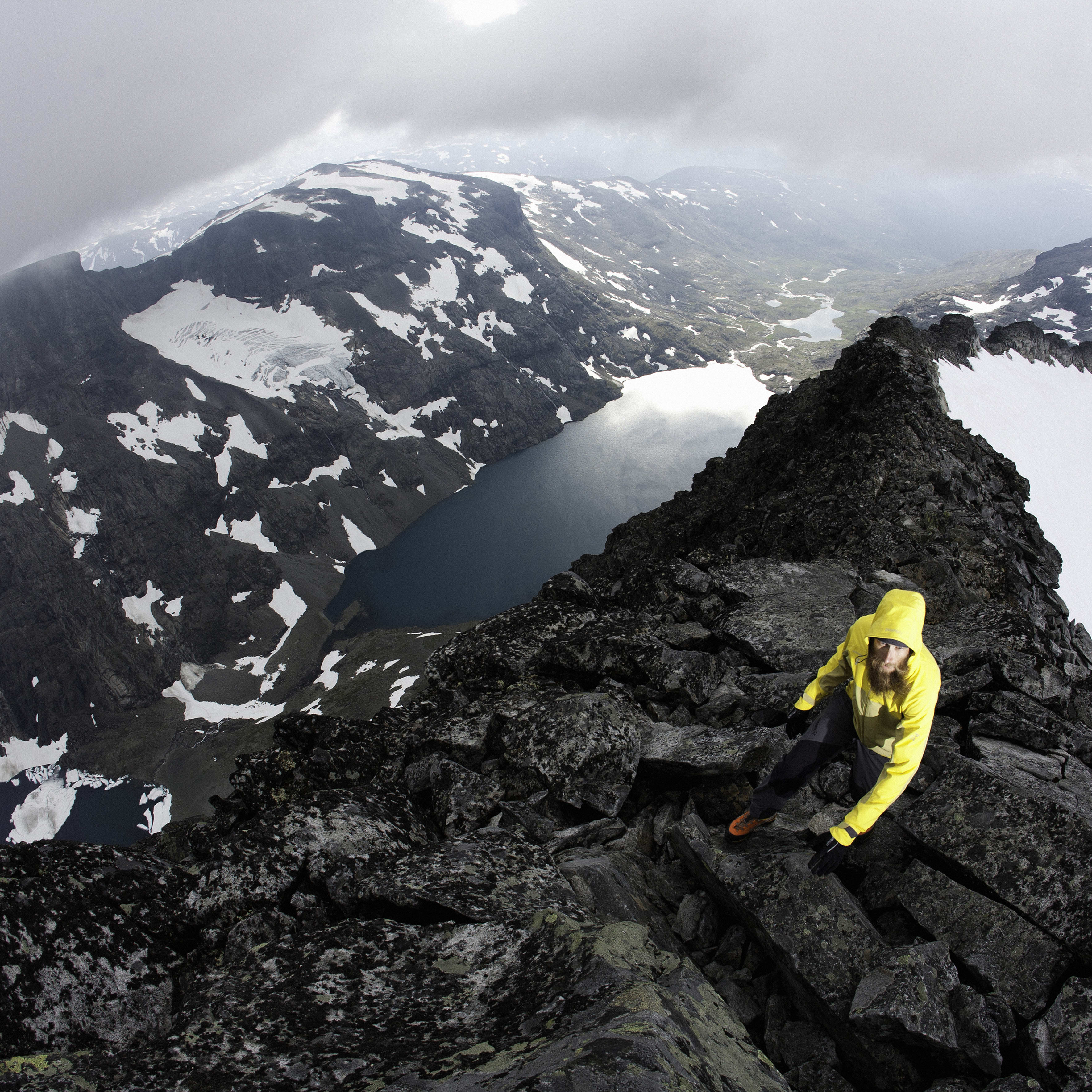 A person in a yellow Norrøna jacket climbs a rocky mountain ridge with snow and a lake visible in the background.