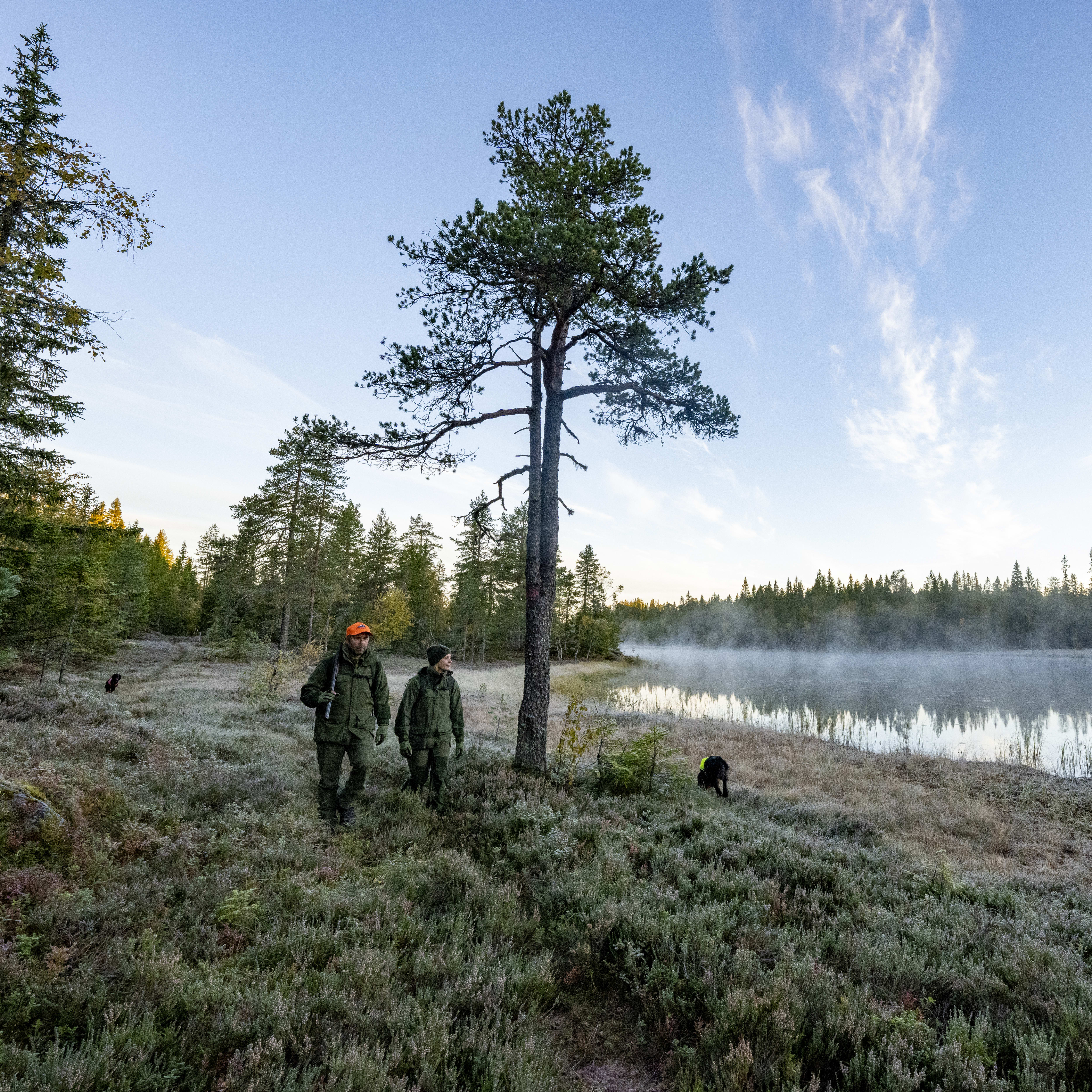 Two people wearing Norrøna gear walk with a dog near a foggy lake in a forest. 
