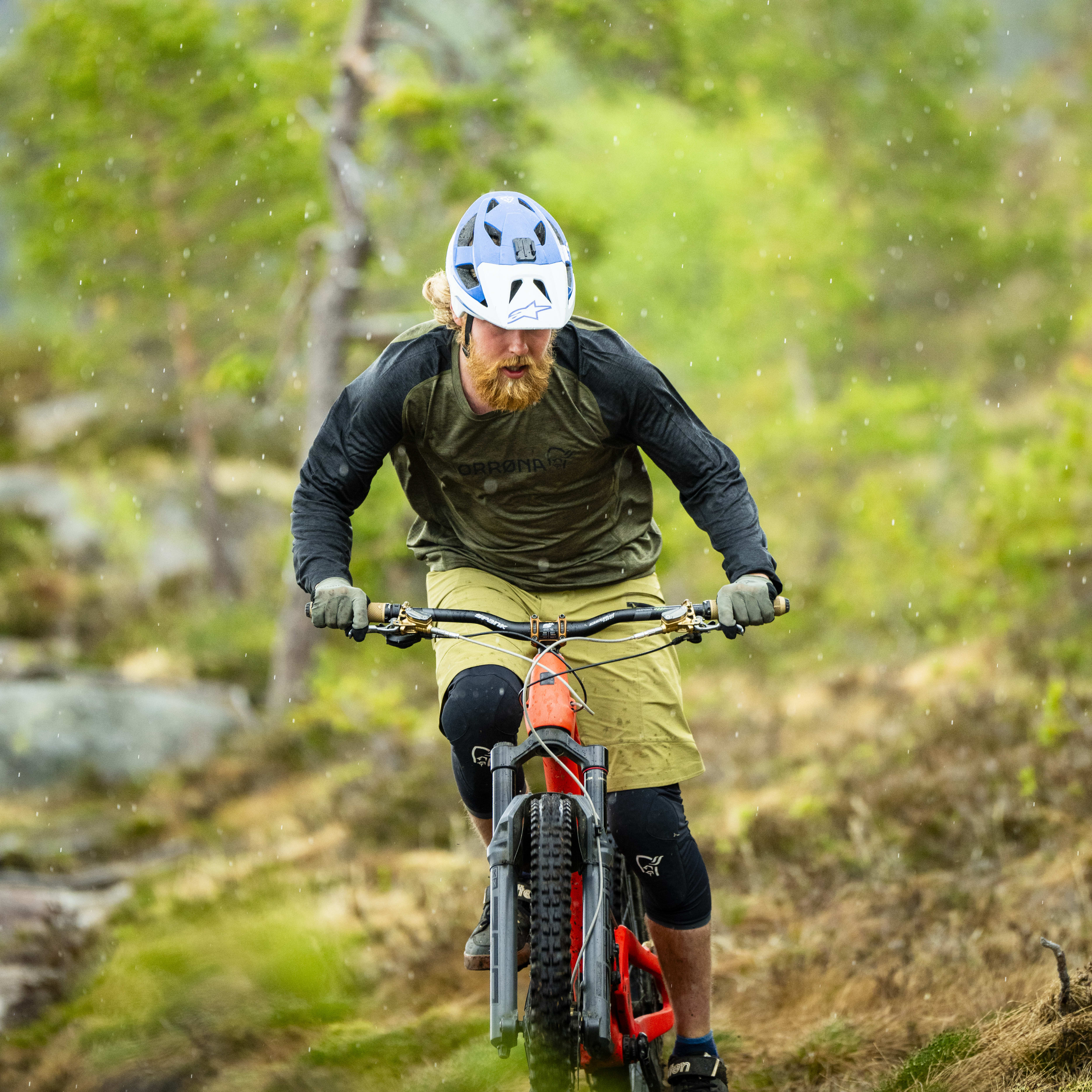 A man wearing a Norrøna shirt rides a mountain bike on a trail in a forest. It's raining.