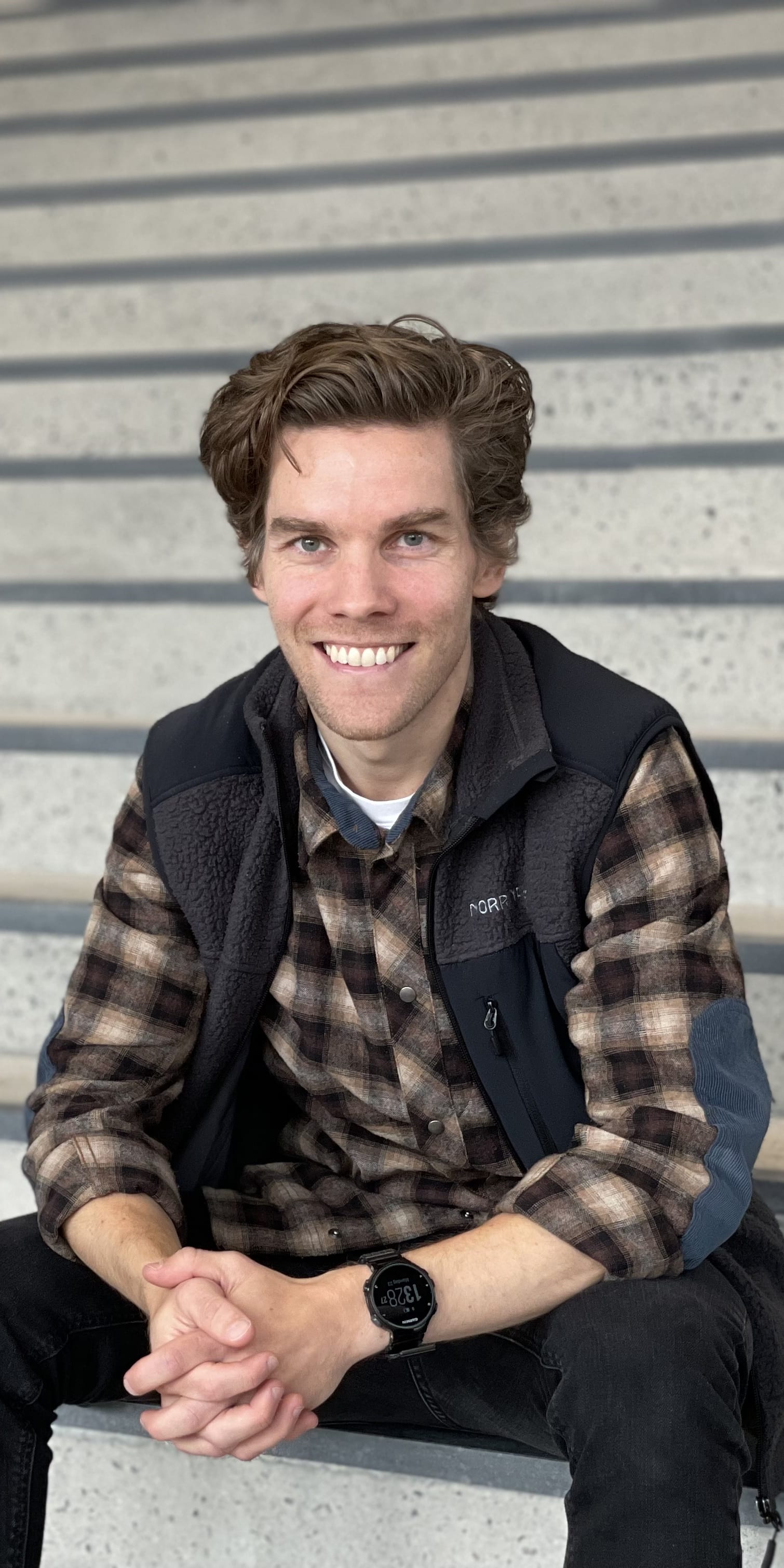A man wearing a brown and black plaid shirt, a black fleece vest, and blue jeans sits on a set of concrete stairs with his hands clasped. He is smiling at the camera. His watch is visible on his left wrist.