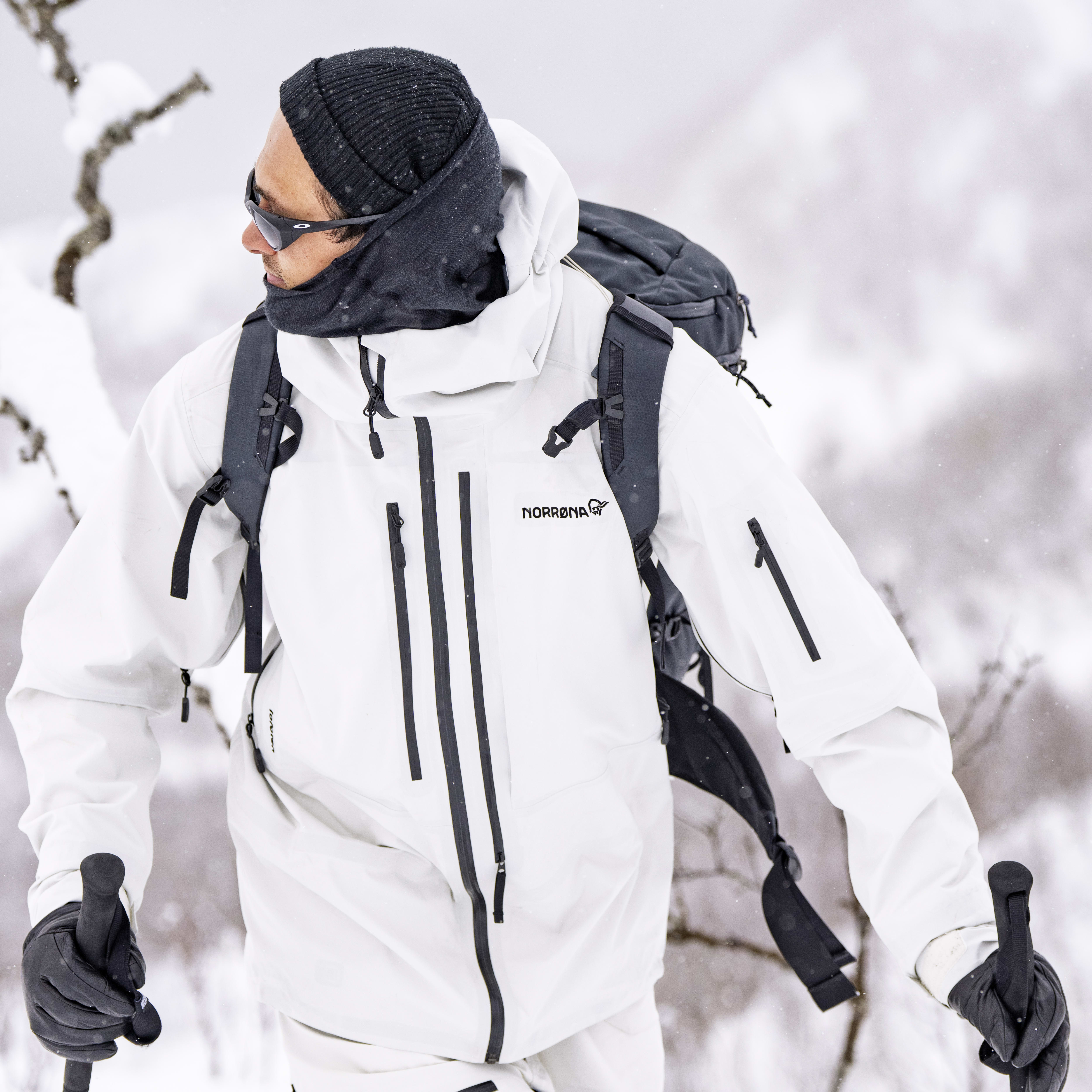 A person wearing a white Norrøna jacket and black pants, a backpack and ski poles, walks through snowy terrain.