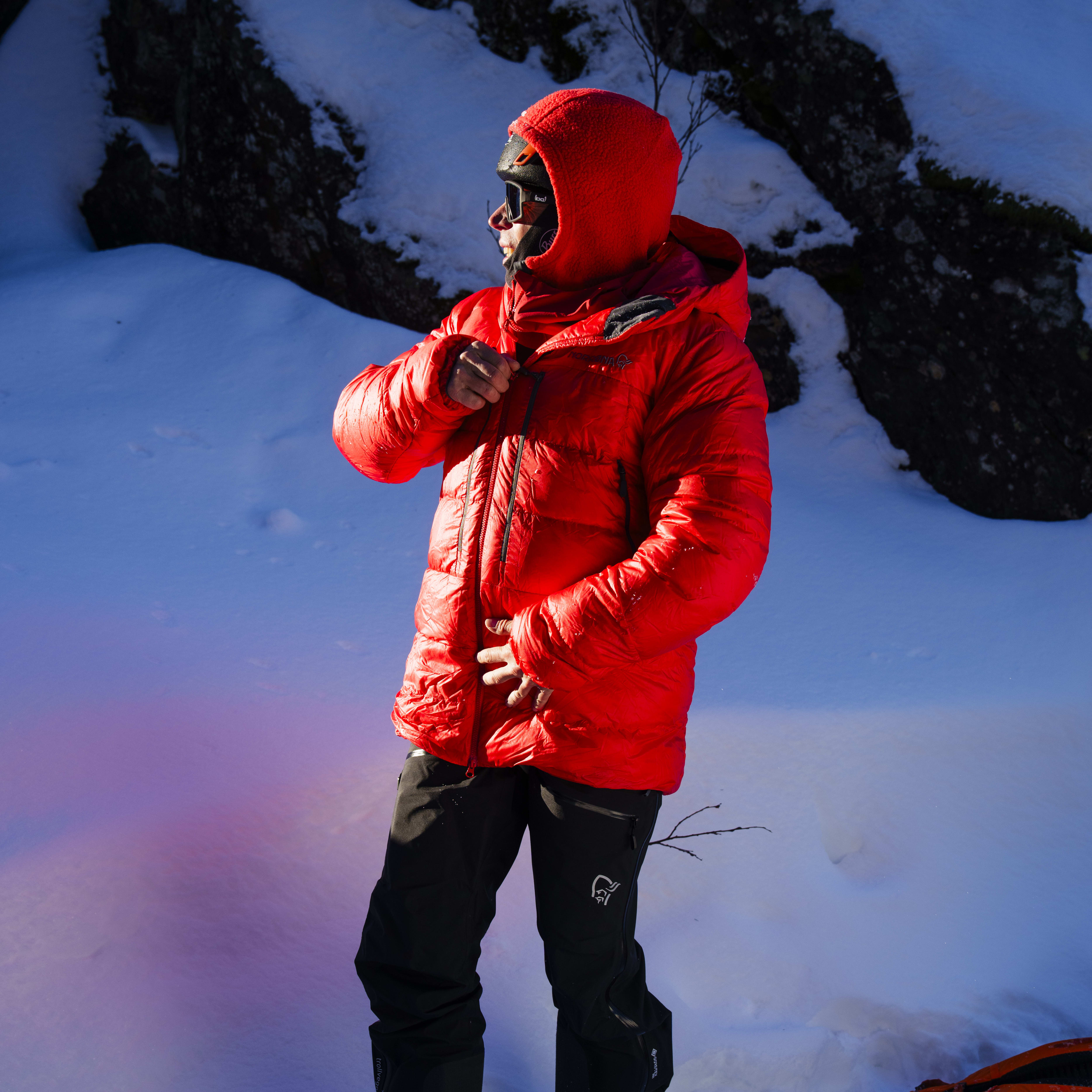 A person wearing a red Norrøna jacket and black pants stands in deep snow. A black glove lies in the snow.