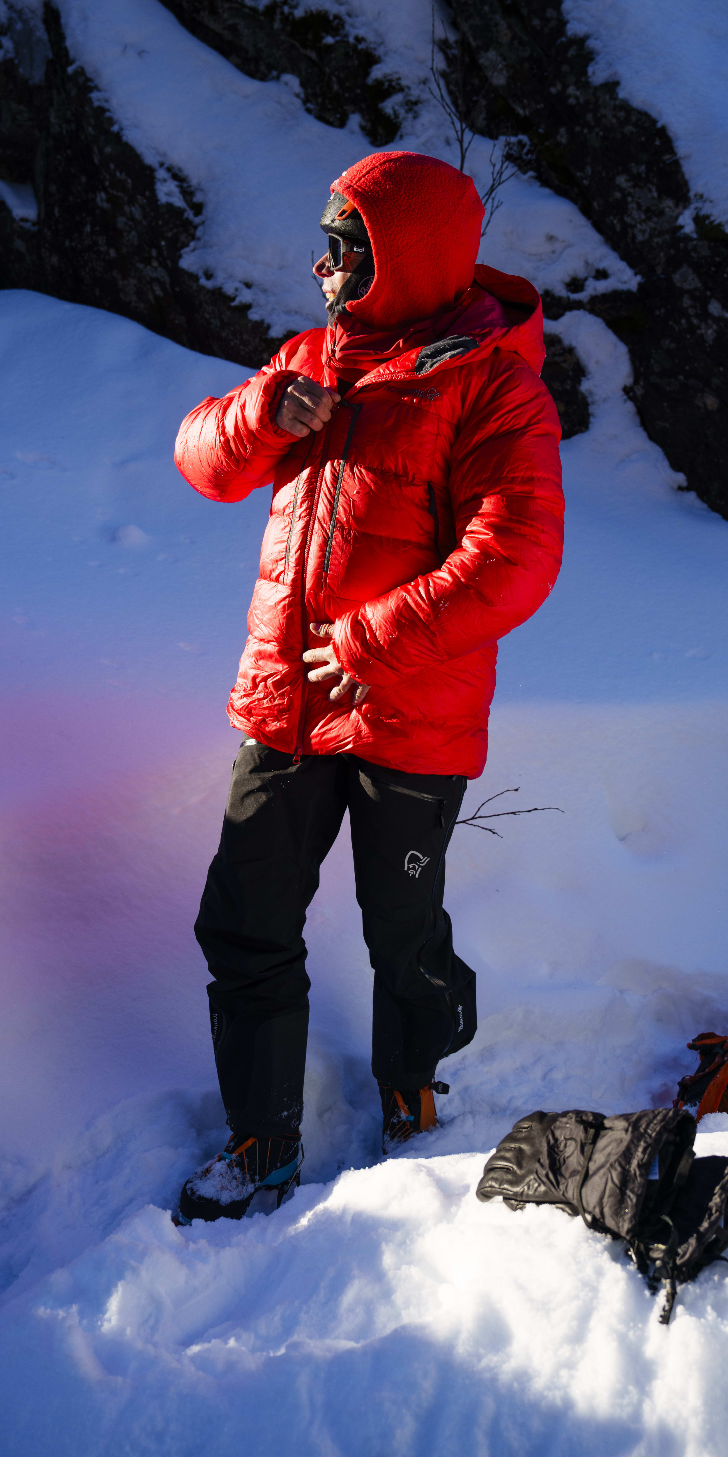 A person wearing a red Norrøna jacket and black pants stands in deep snow.  A black glove lies in the snow.  
