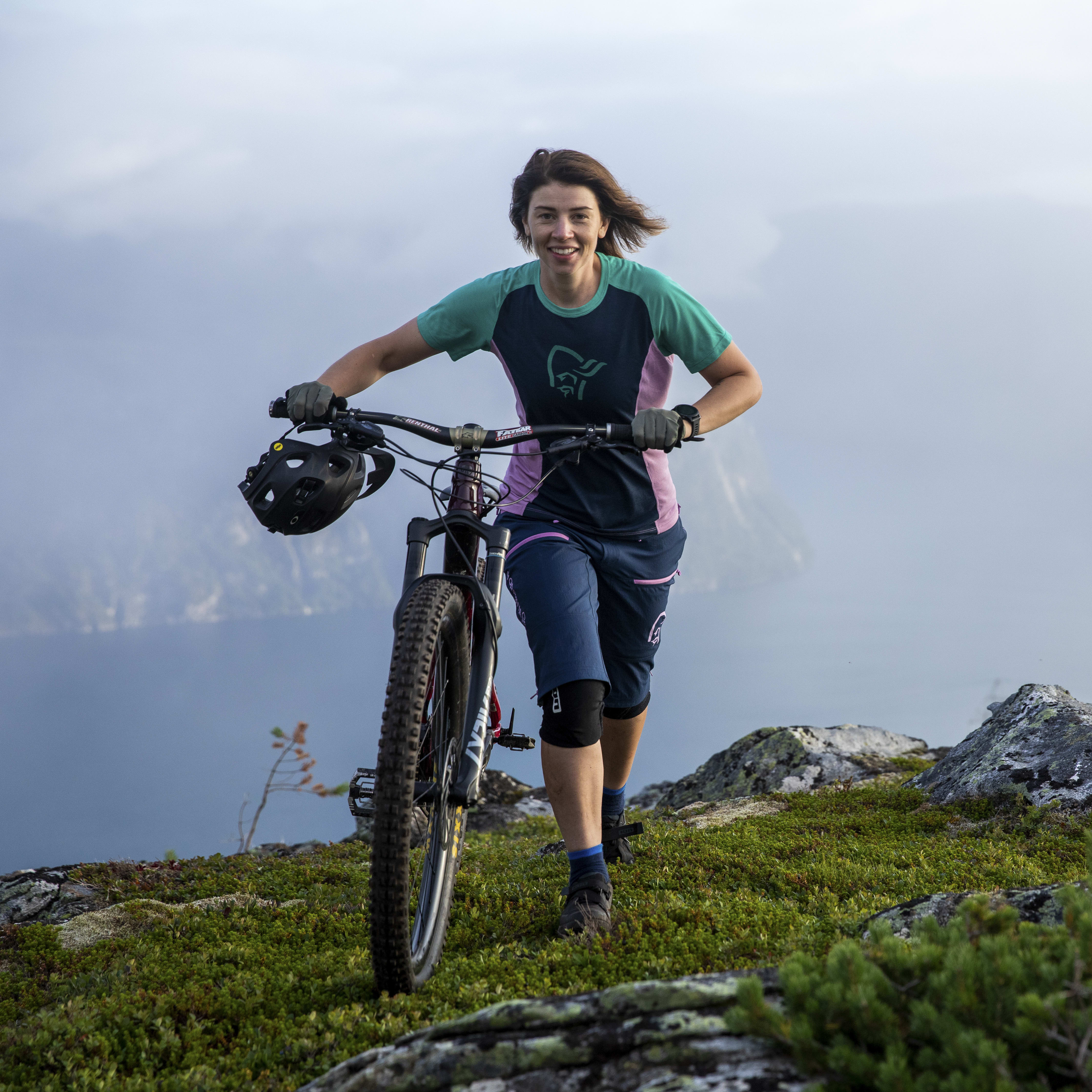 Woman on a mountaintop walks with a mountain bike, wearing a Norrøna shirt and shorts. Ocean and misty mountains are visible in the background.