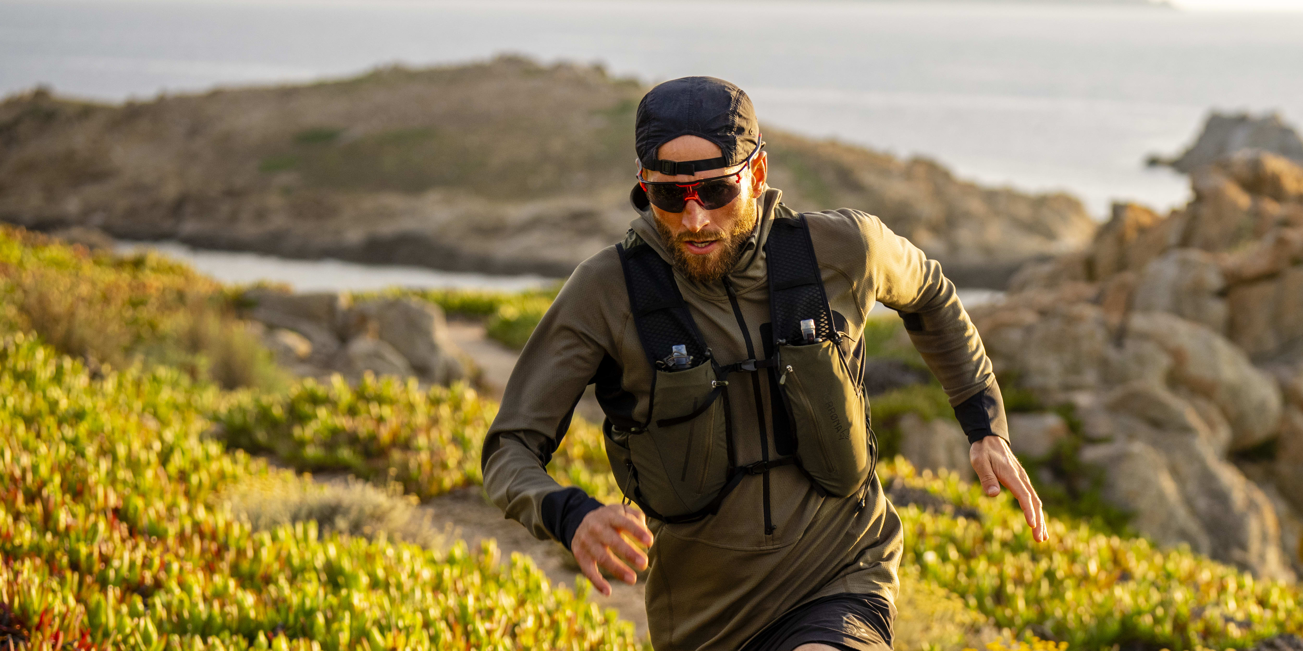 A man wearing a Norrøna running vest and shorts runs along a coastal trail.