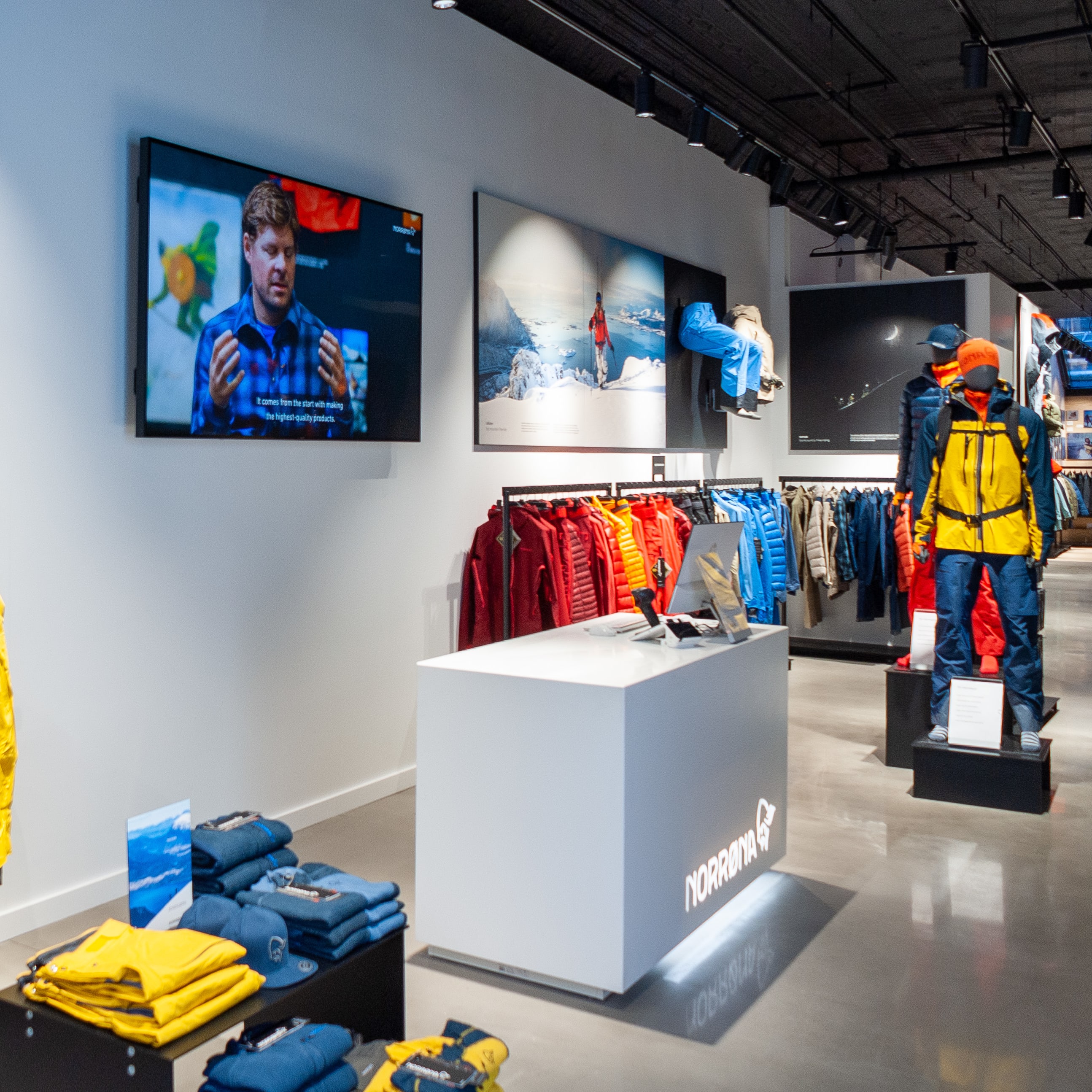 Norrøna store interior with display of men's outerwear and a mannequin wearing a yellow jacket and blue pants.  The store has a modern design with white walls and a white counter with the Norrøna logo. 
