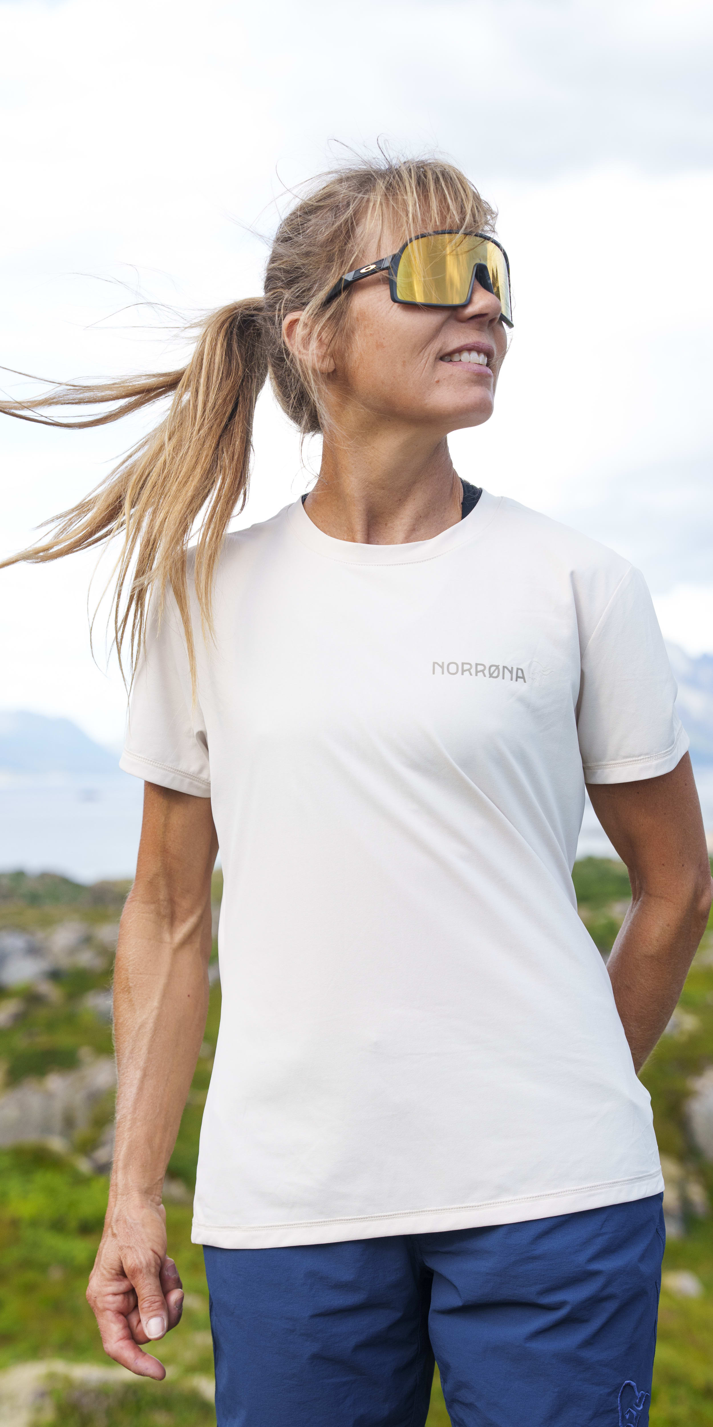 Woman in Norrøna t-shirt and shorts wearing sunglasses, looking to the side outdoors with mountains in the background.
