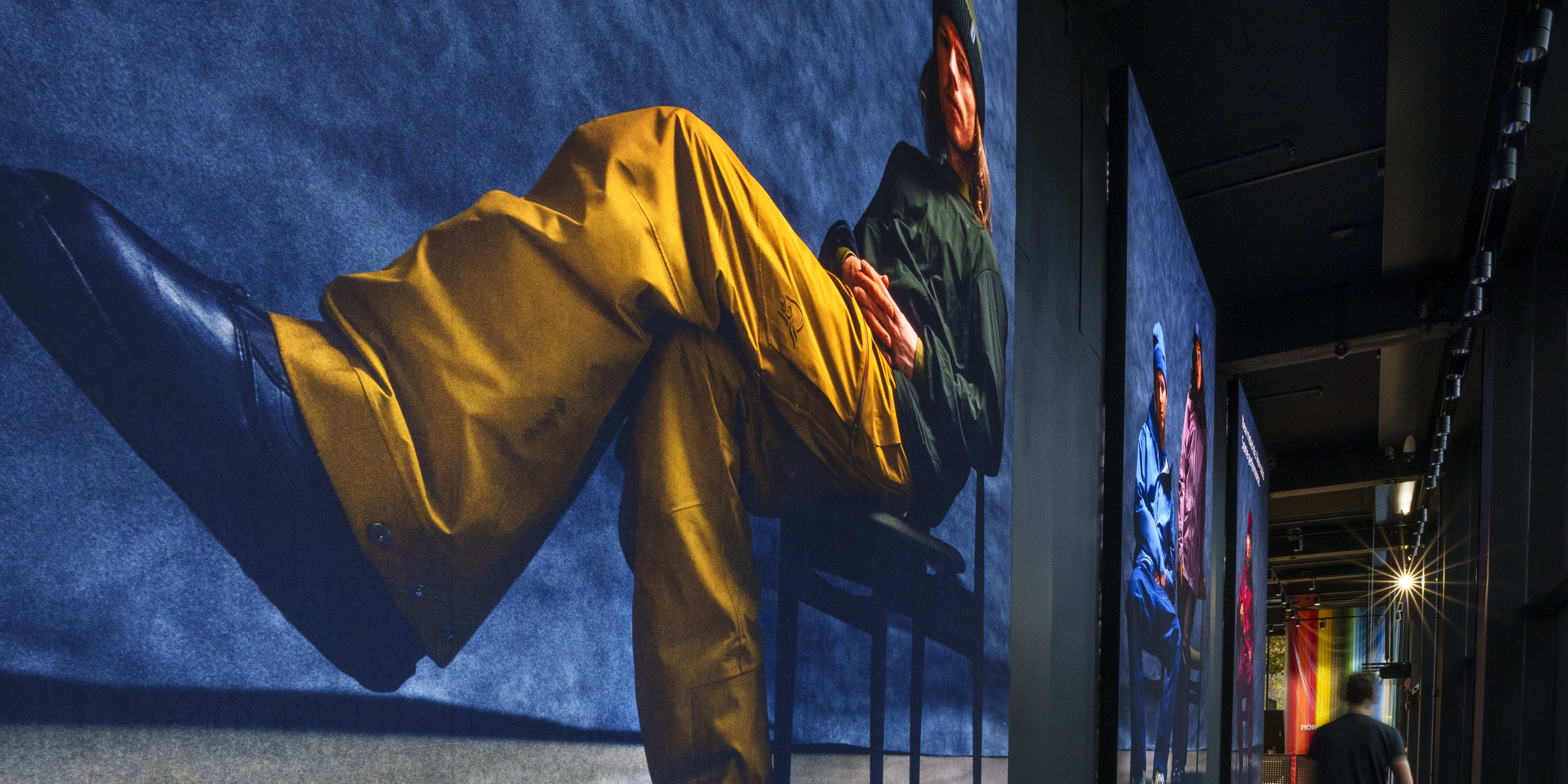 A person wearing a green jacket and a hat, yellow pants, and blue shoes sits on a chair.  They are posed in a way that makes it appear as though they are facing the right side of the image.  The person is in front of a large blue background that has the words "The new Gore-Tex fabric with an innovative ePE membrane comes with all well-known benefits of a Gore-Tex product." written on it.  The image also includes the GORE-TEX logo. There is a person walking away from the image in the background.  All clothing is from Norrøna. 
