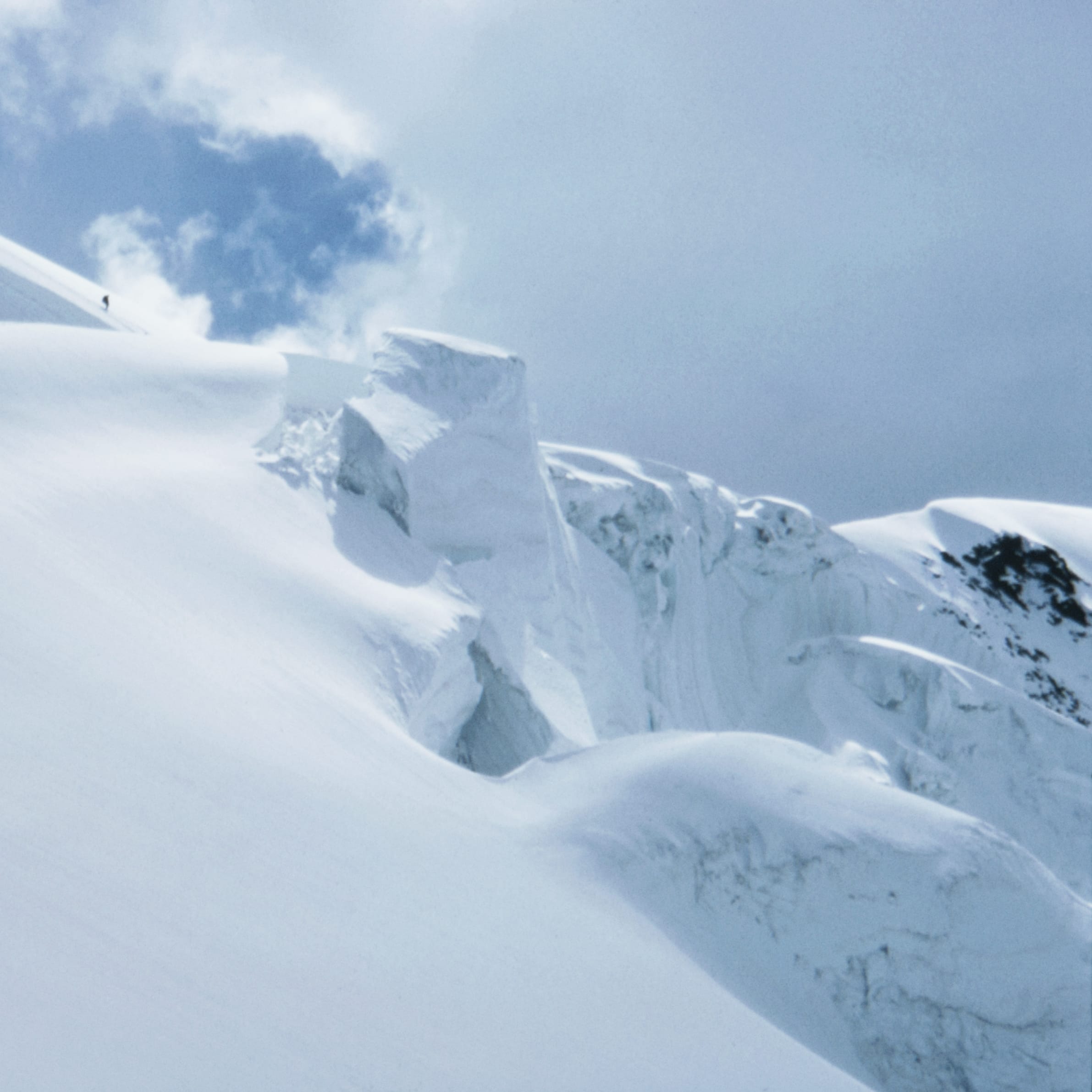 A snowy mountain peak with a person climbing in the distance. The sky is mostly blue with some white clouds.