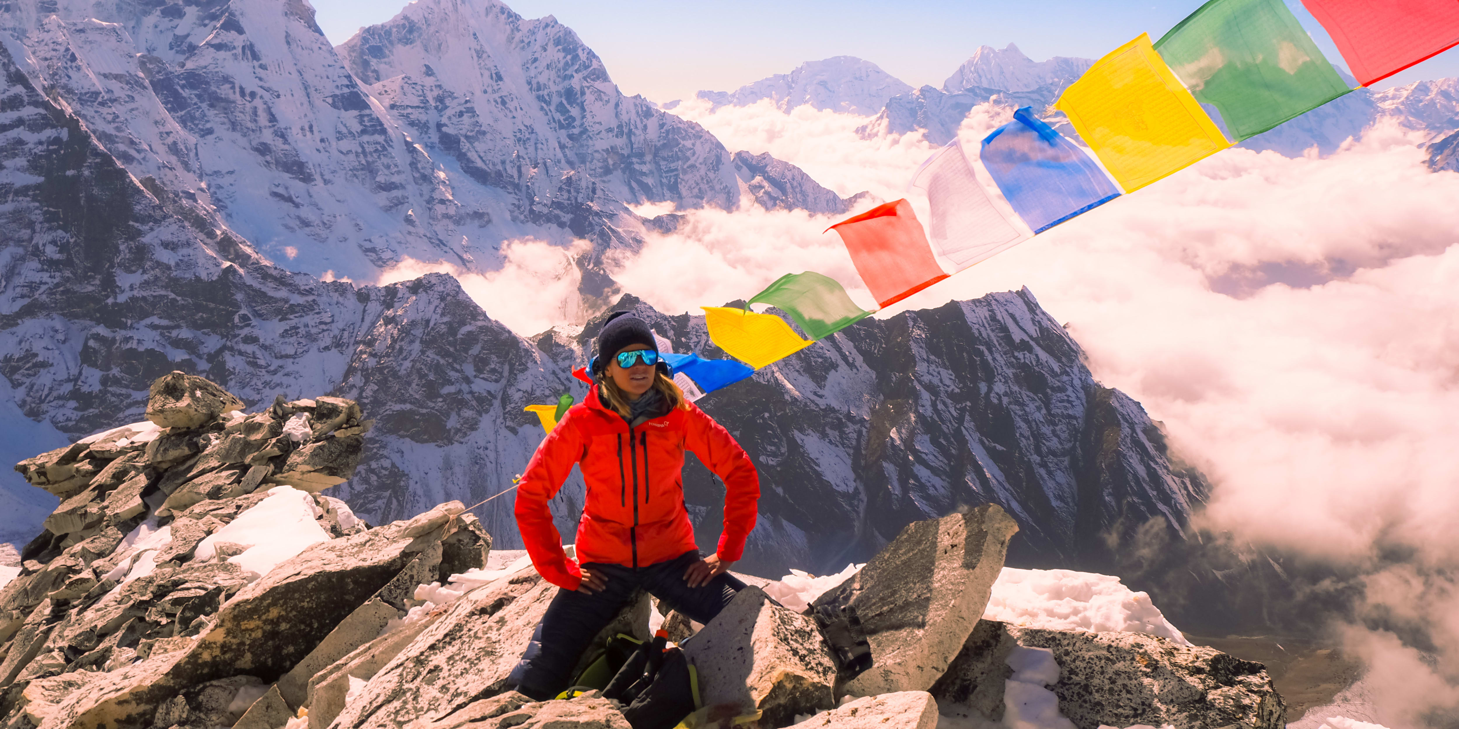 Elisa Røtterud in Himalaya on the top of a mountain with typical Himalayaan flags in all different colors. The sun is shining and the mountains are covered in a light snowpack. 
