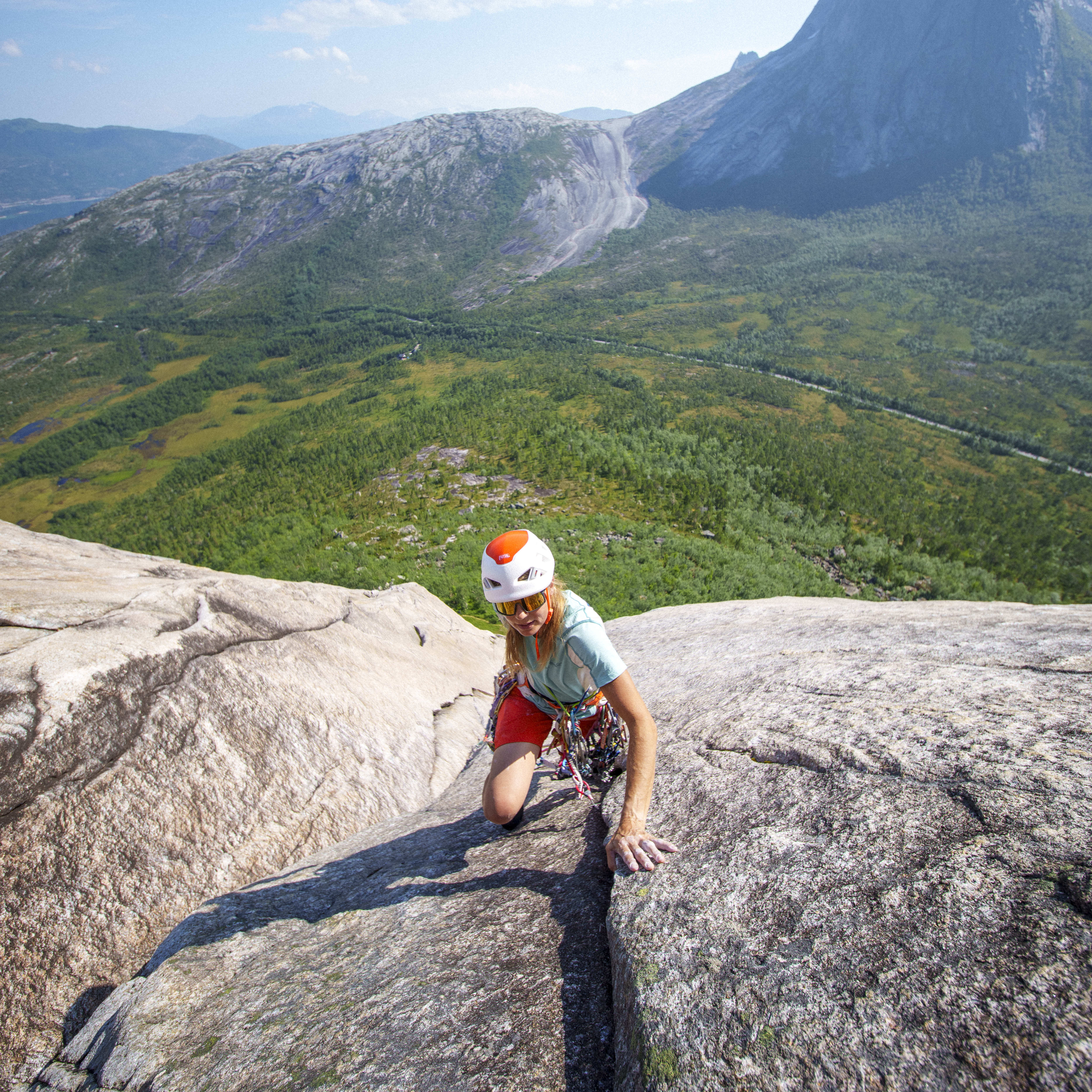 A climber with a helmet ascends a rock face. They wear Norrøna shorts and gear, with a valley and mountain visible in the background.
