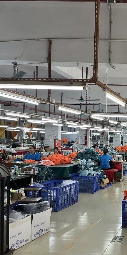 A large factory room with many people working at sewing machines. They are all wearing blue shirts and working on orange fabric. There are many blue plastic crates filled with fabric around the room.