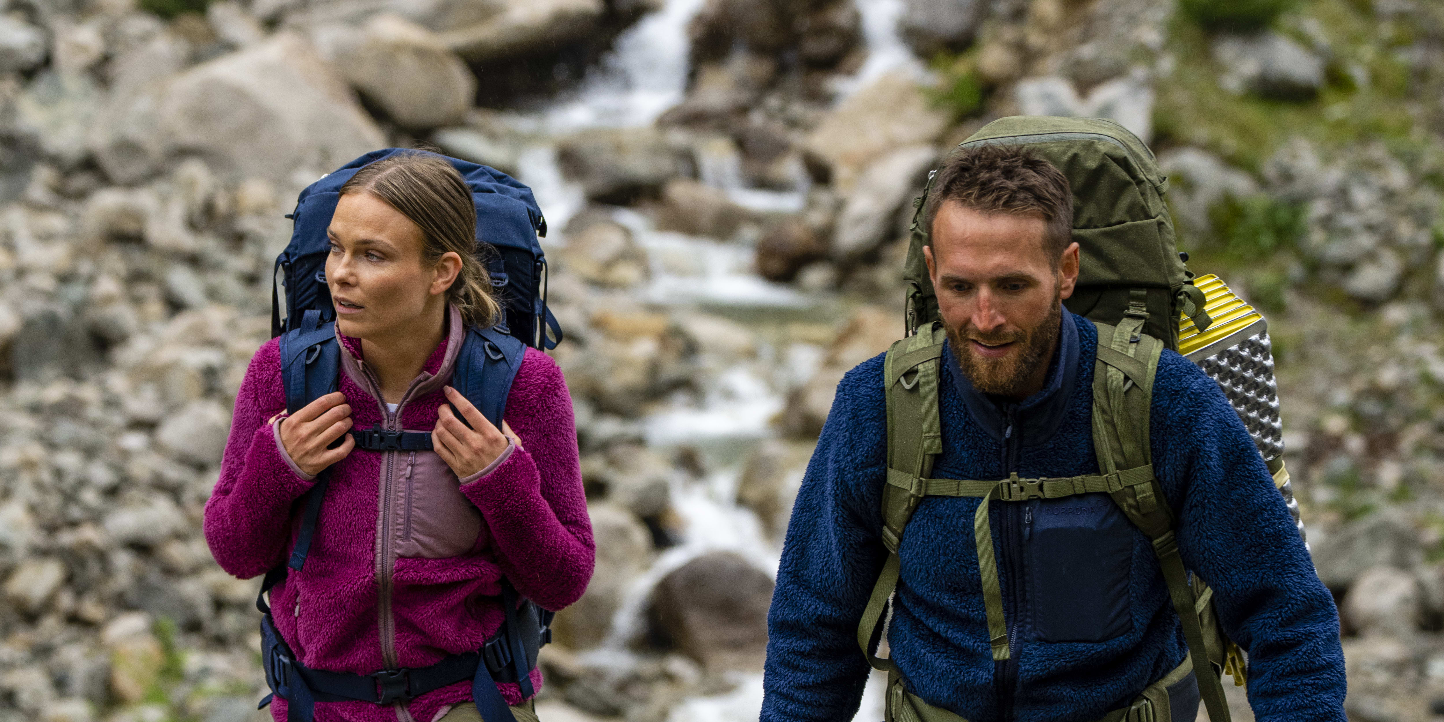 Two people wearing Norrøna gear hike through a rocky landscape with a small stream.