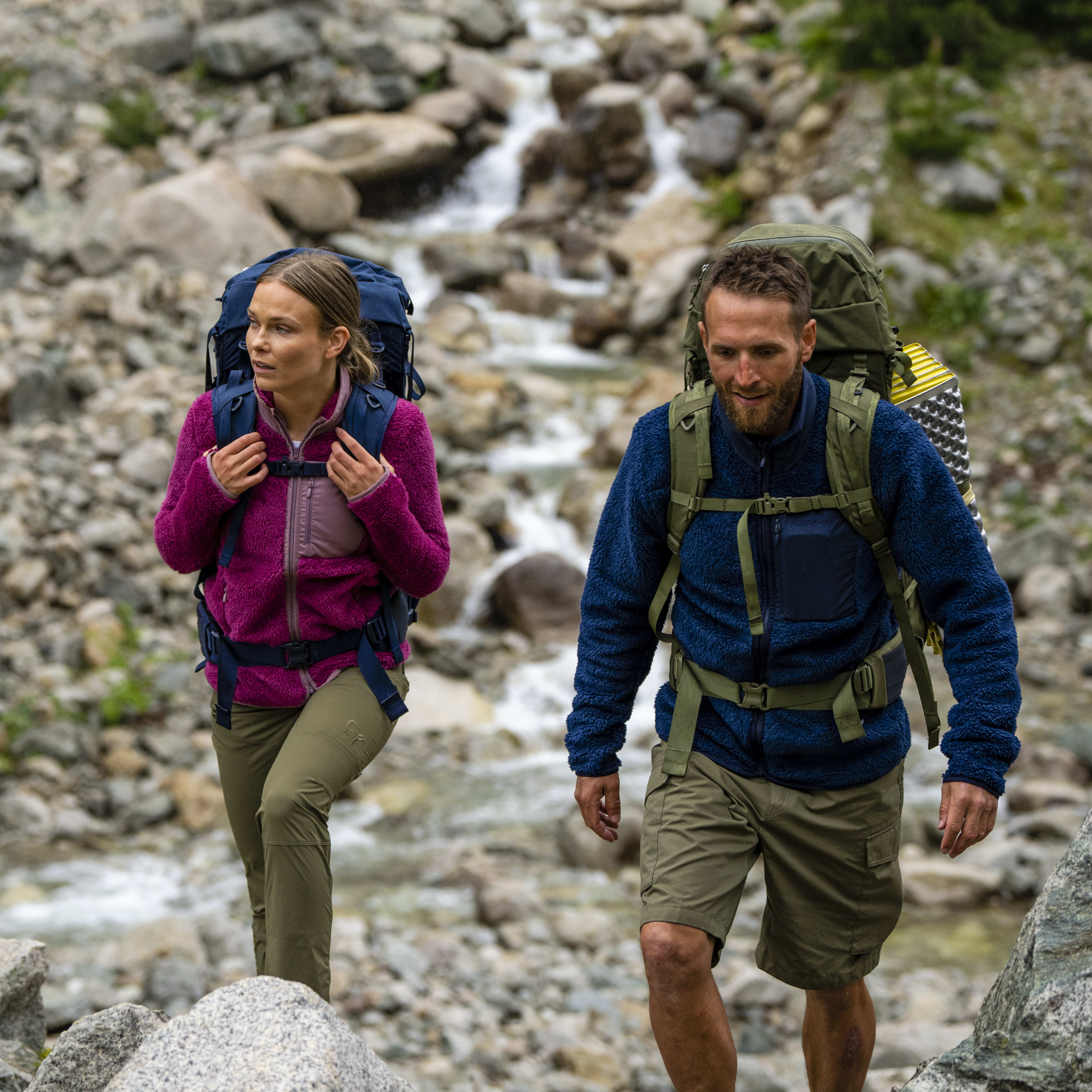 Two people wearing Norrøna gear hike through a rocky landscape with a small stream.