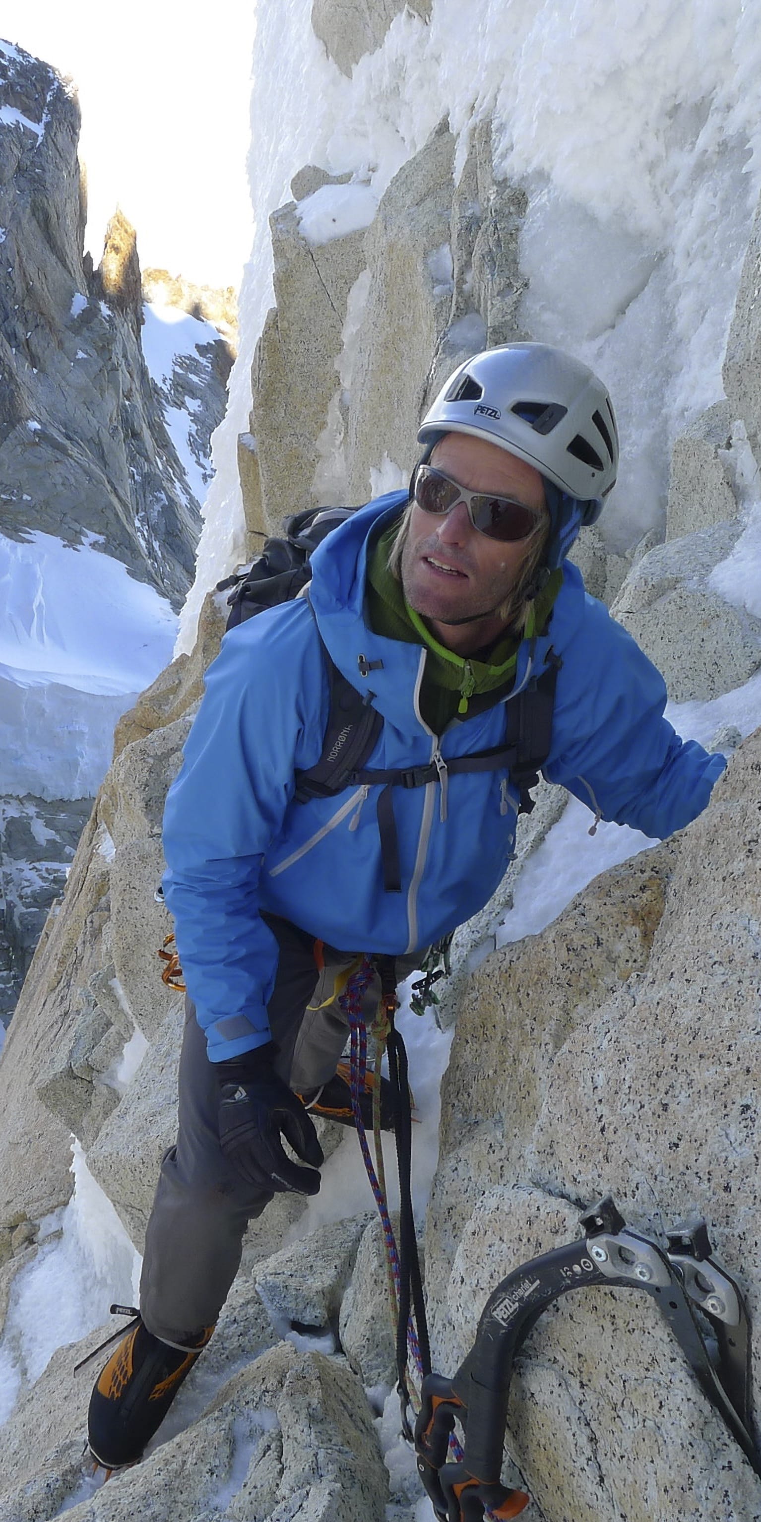 A rock climber wearing a helmet and a blue jacket is climbing a snowy mountain face.  The climber is using a technical ice axe and has a rope attached to their harness.  The background shows a large snowy mountain face.