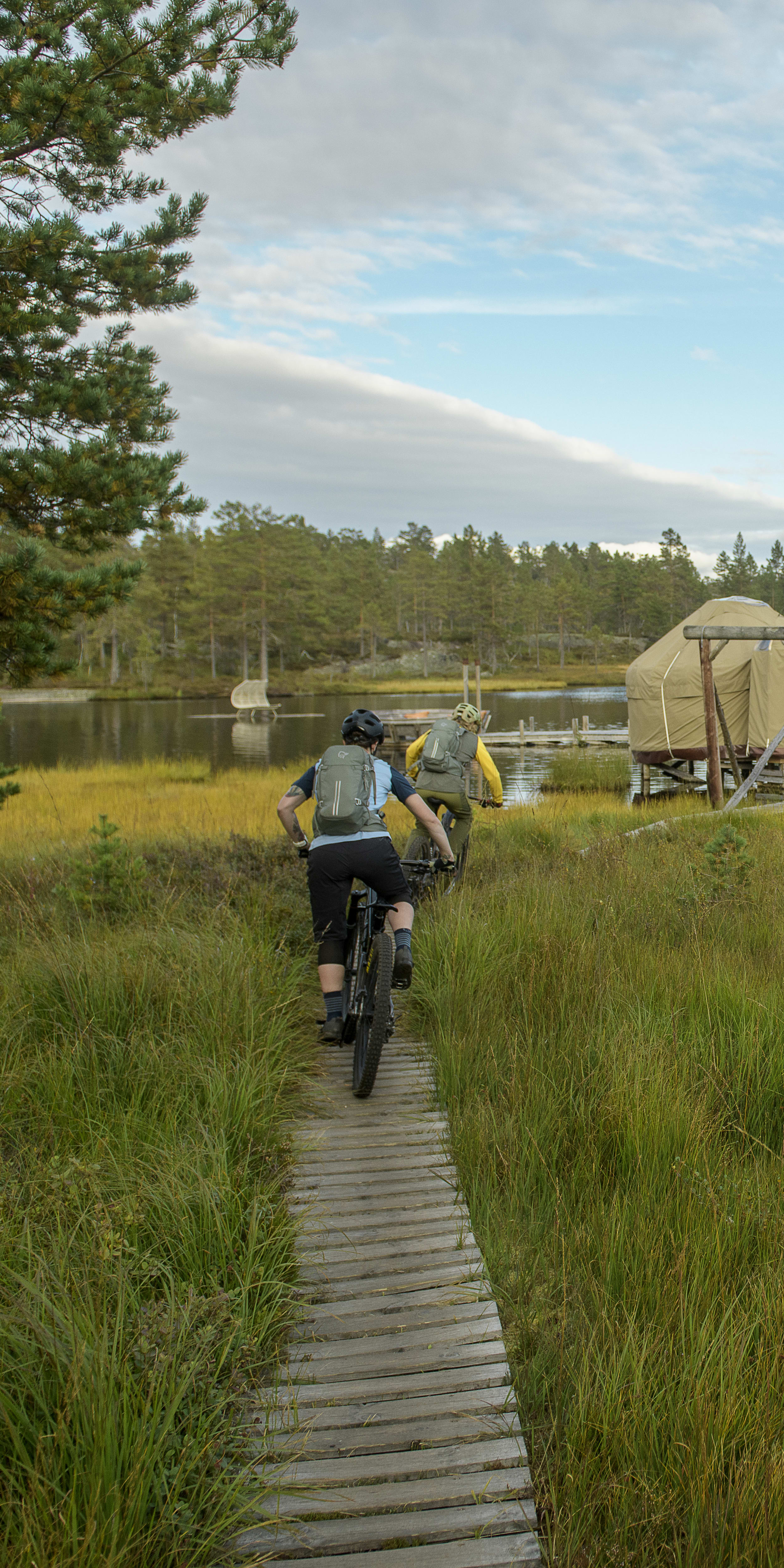 Two people riding mountain bikes on a wooden path in a grassy field. They are passing by a lake and some cabins. One biker is wearing a Norrøna backpack. 
