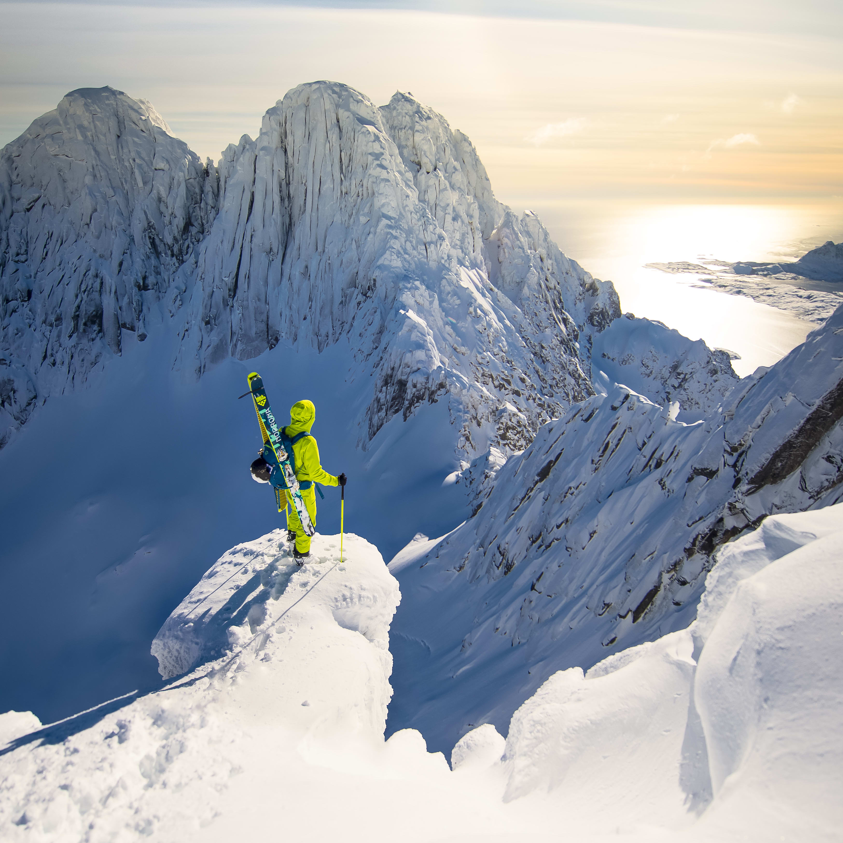 A person in yellow Norrøna gear stands on a snow-covered mountain peak with a pair of skis and ski poles.  A vast expanse of snow-capped mountains and a glistening body of water stretch out below. 
