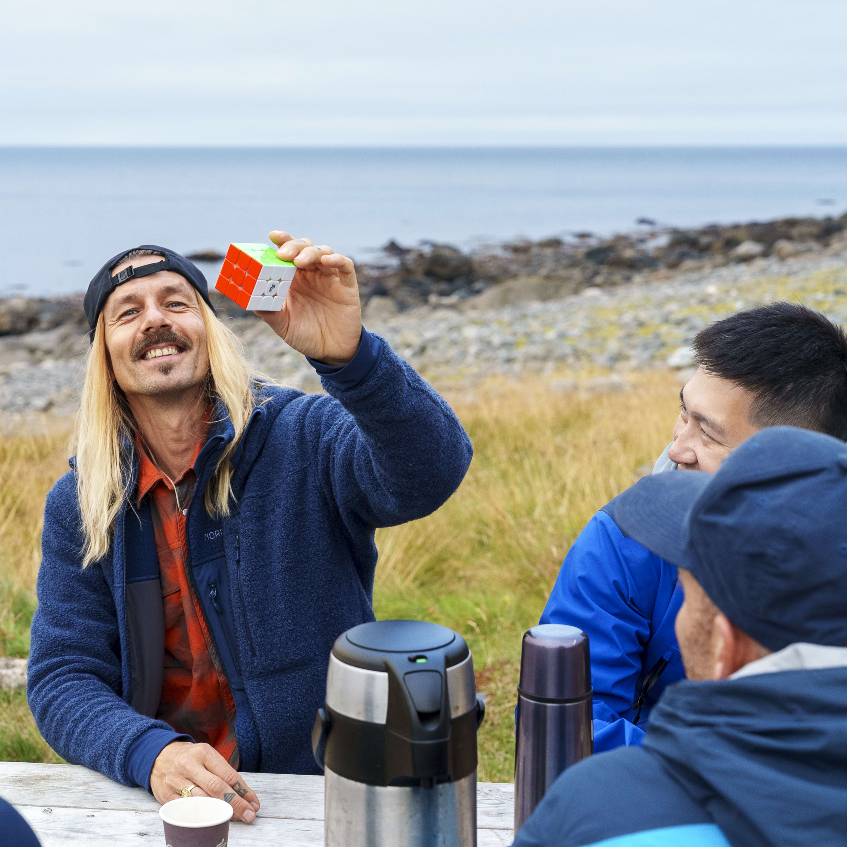 A man in a blue Norrøna jacket is holding a Rubik's Cube while smiling at the camera. He is with two other men.