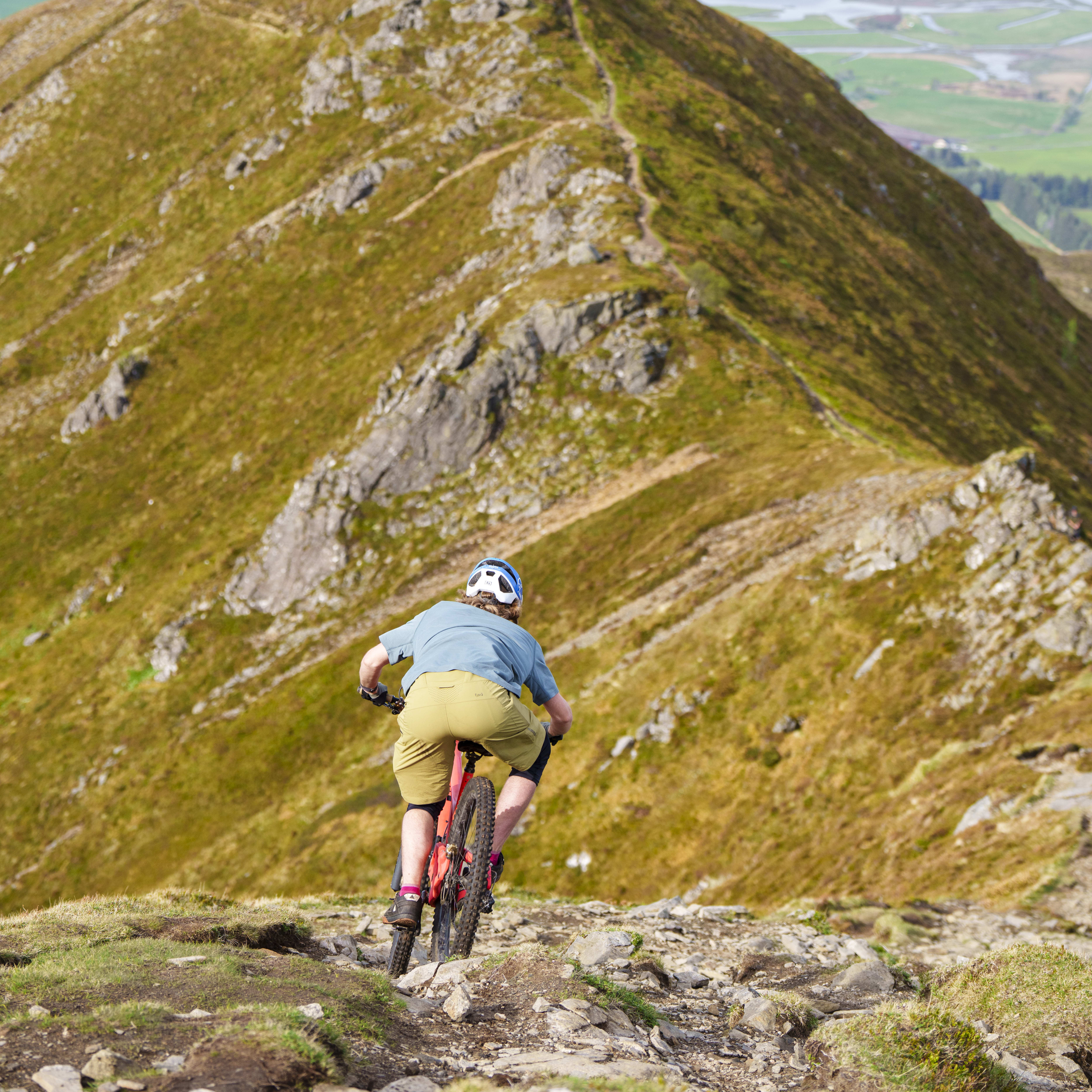 Person riding a mountain bike on a rocky trail with a view of a fjord in the distance. They are wearing a Norrøna shirt and pants.