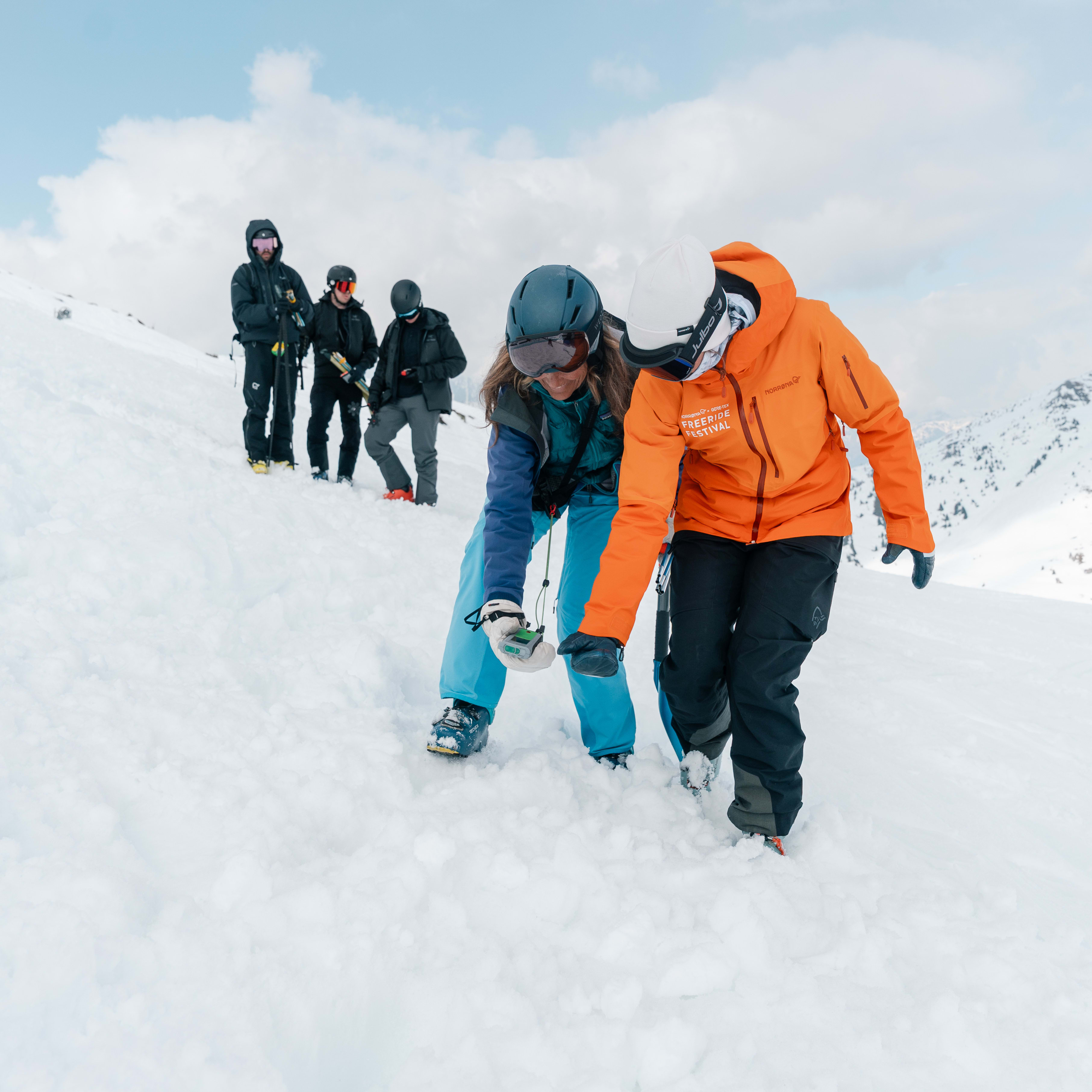 Skiers in Norrøna outerwear practice an avalanche transceiver search on a snowy mountain slope.
