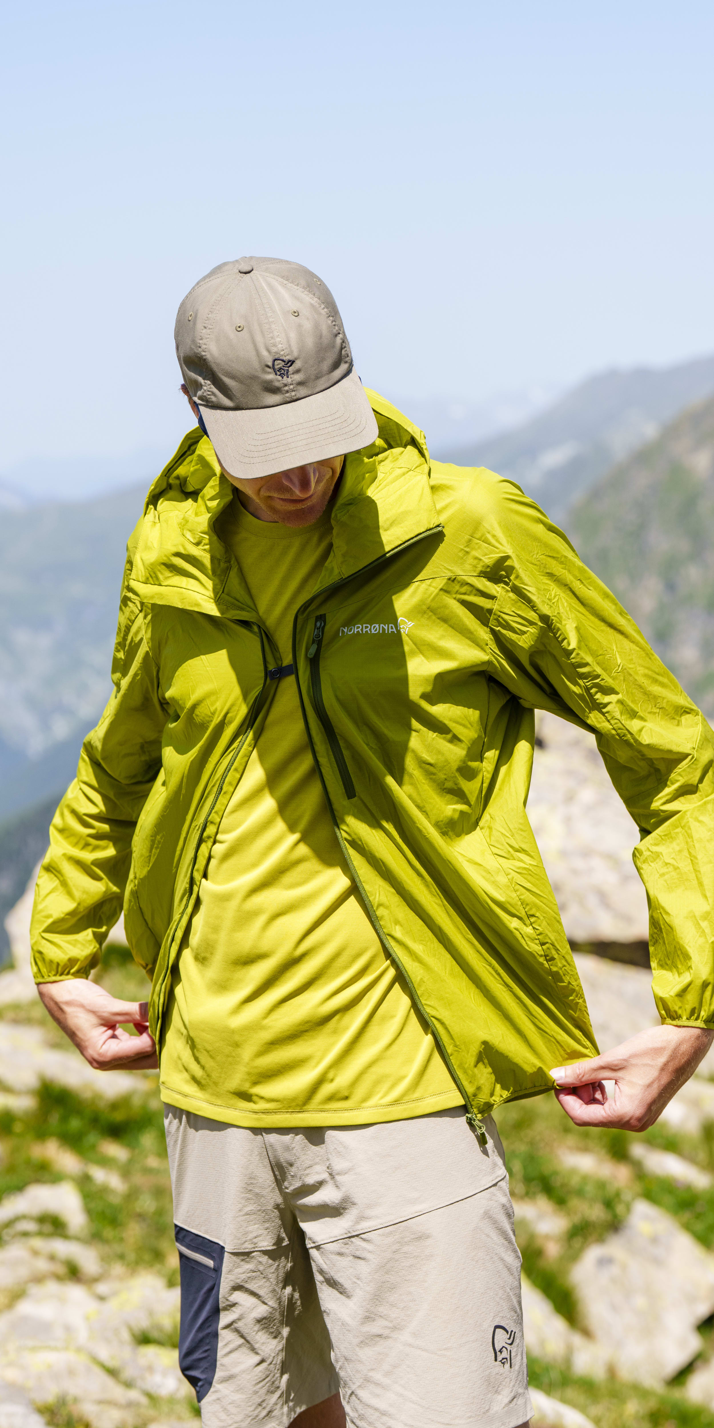 A hiker in a green Norrøna jacket and hat, standing on a rocky mountain with blurred mountains in the background.