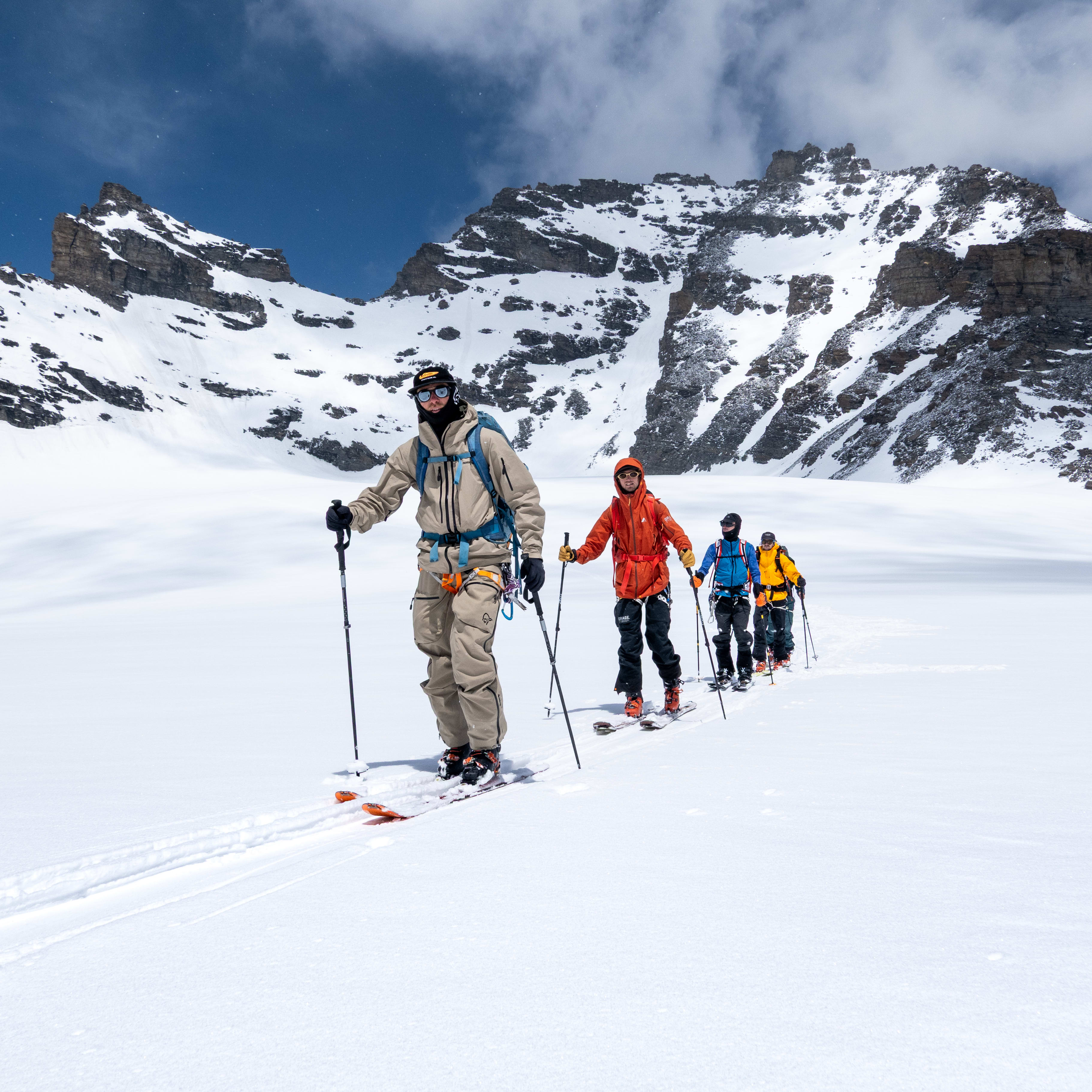 A group of five people ski touring on a glacier, with a large mountain in the background. The first person wears a Norrøna jacket and pants.