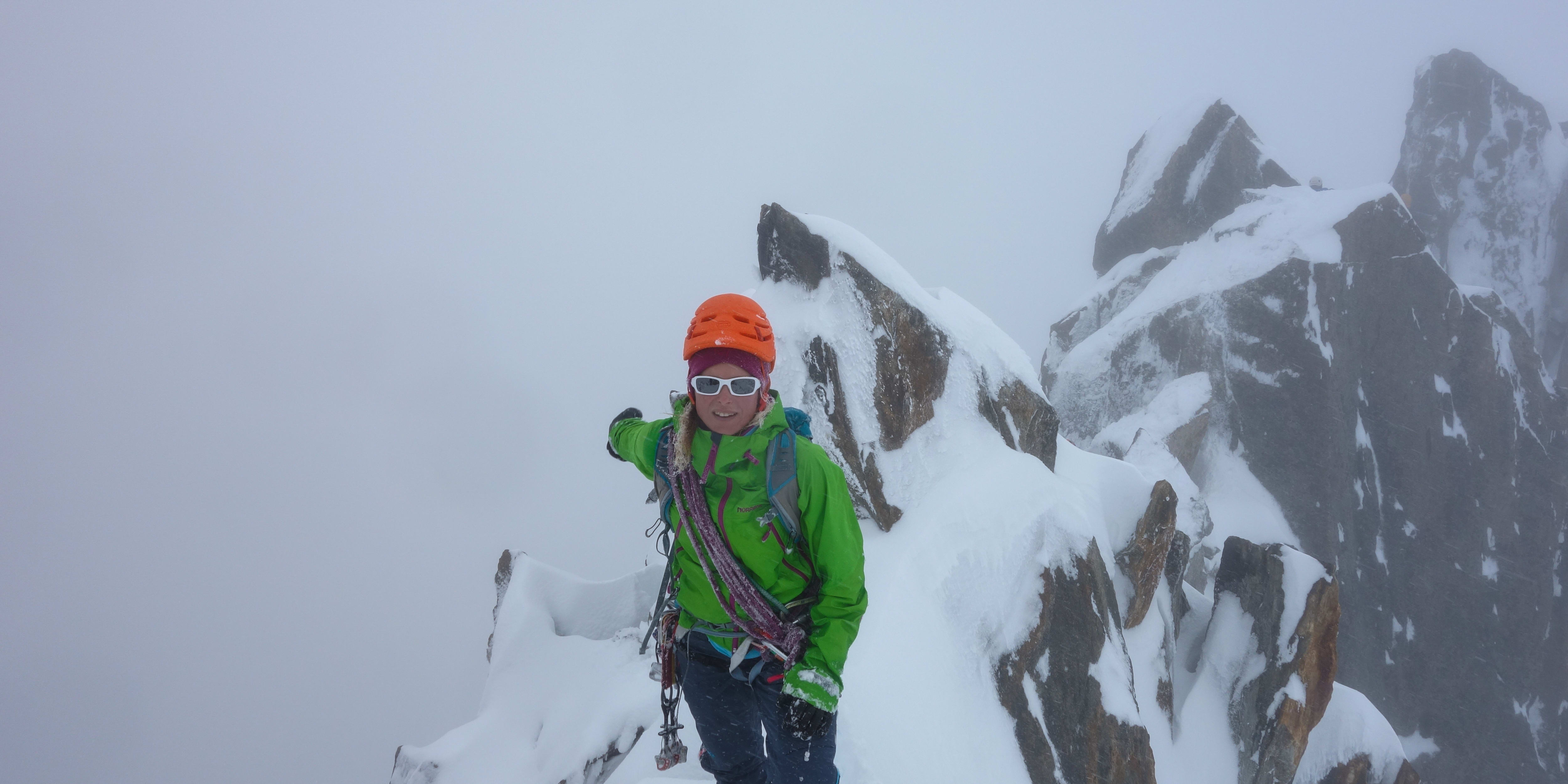 A smiling climber in Norrøna jacket, orange helmet, and sunglasses stands on a snowy, rocky peak with dense fog.