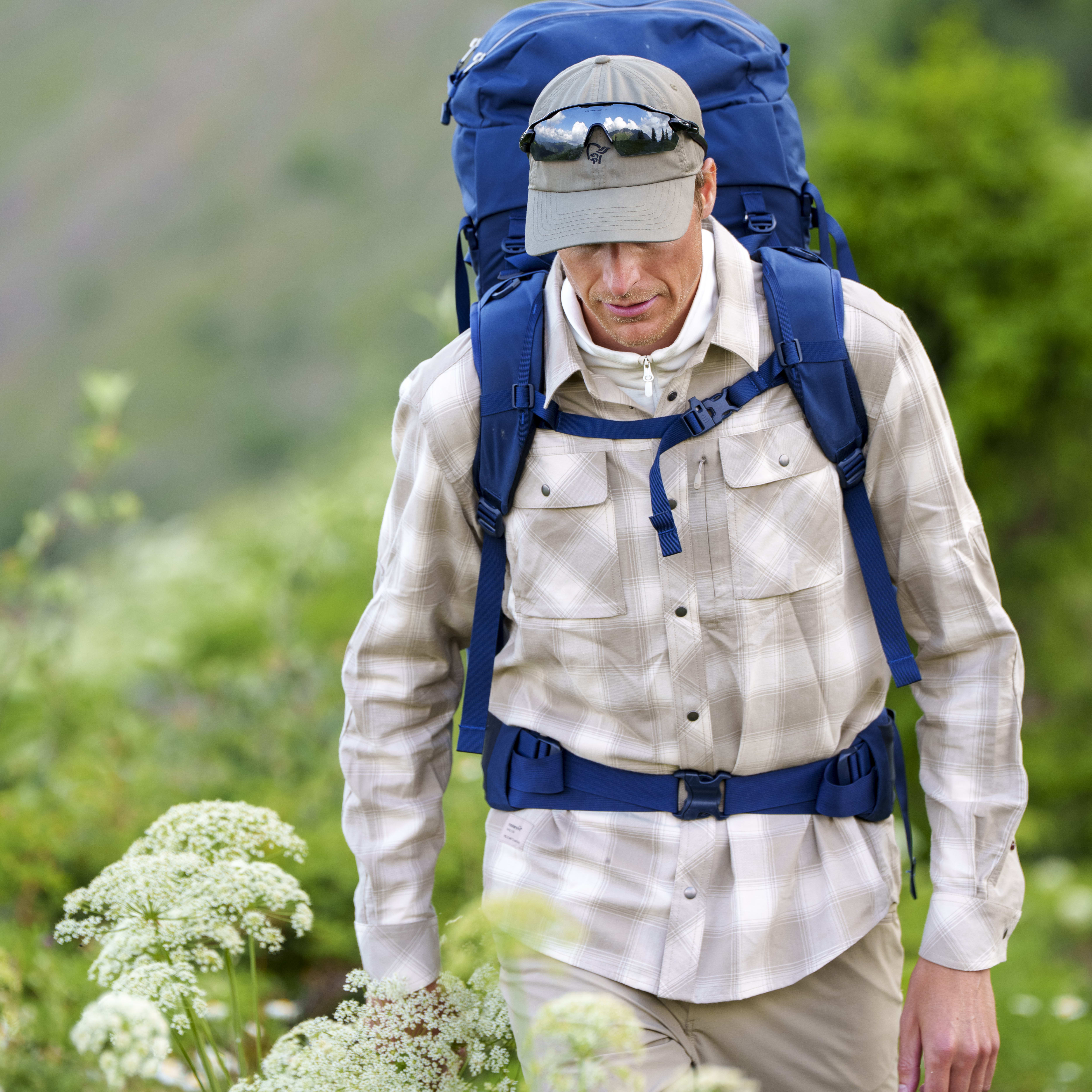 A hiker with a large blue backpack wearing a Norrøna shirt and cap, with sunglasses, walks through a field of wildflowers.