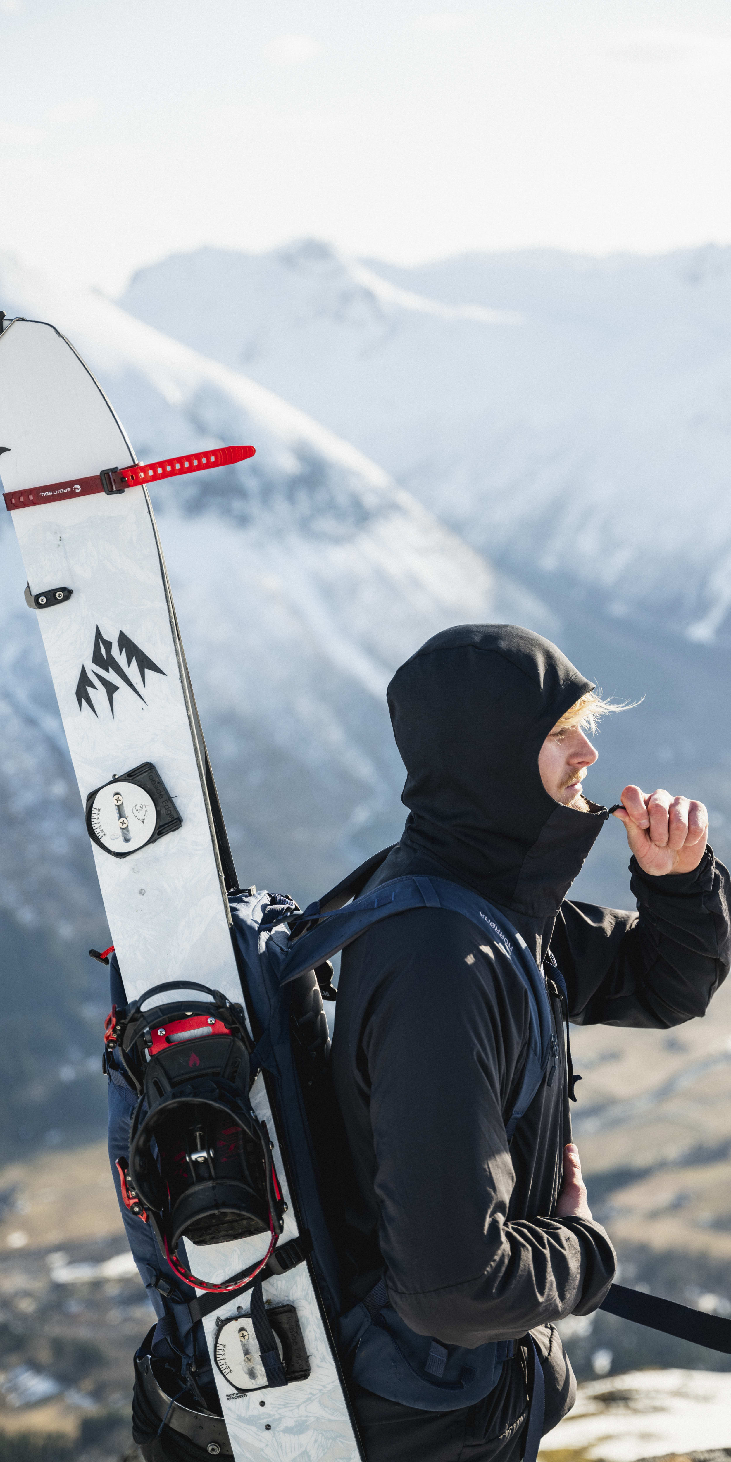 A person wearing a Norrøna jacket stands on a snowy mountain with a splitboard strapped to their back.