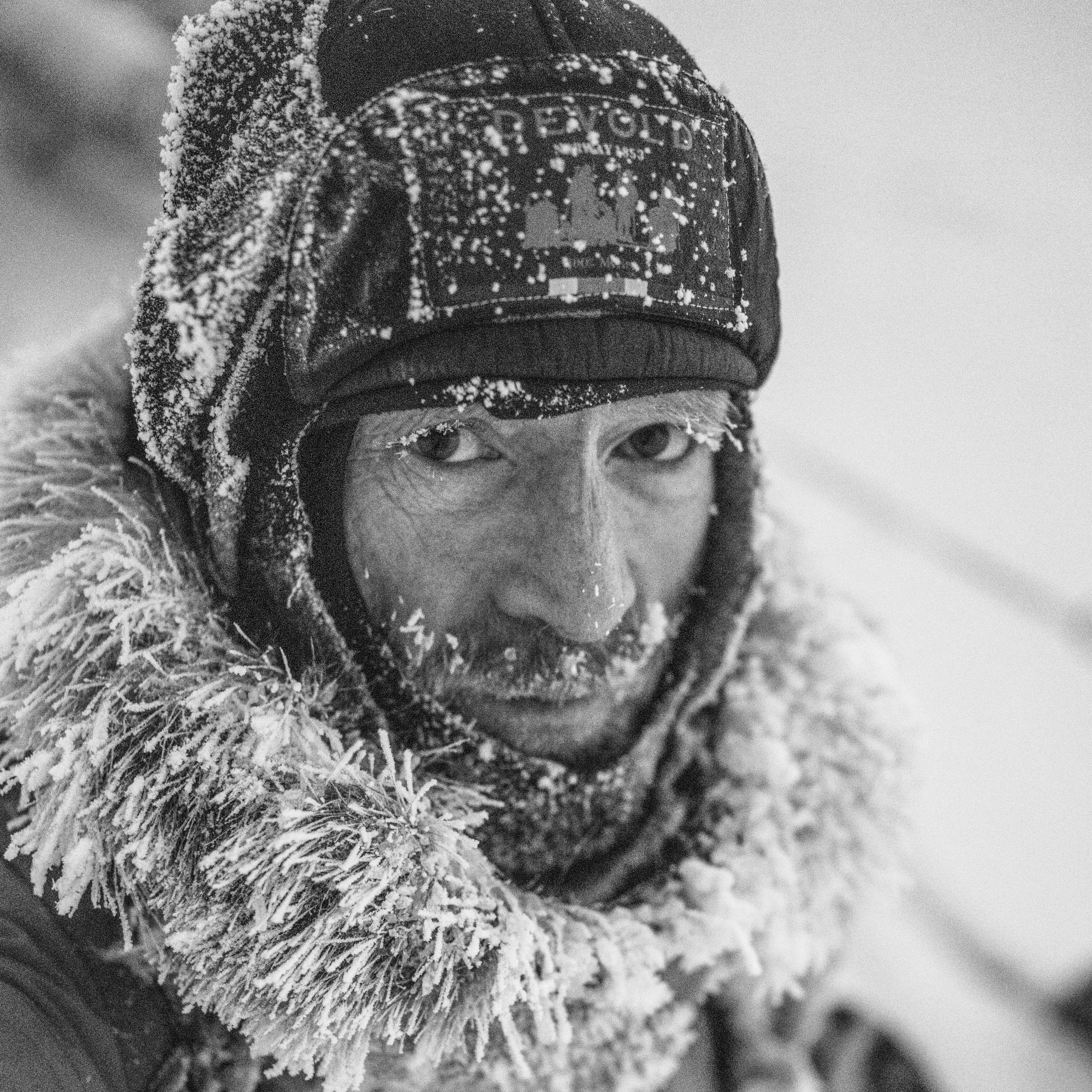 A man wearing a Norrøna hat and jacket is covered in snow and ice. The cold weather creates a stark and textured image.