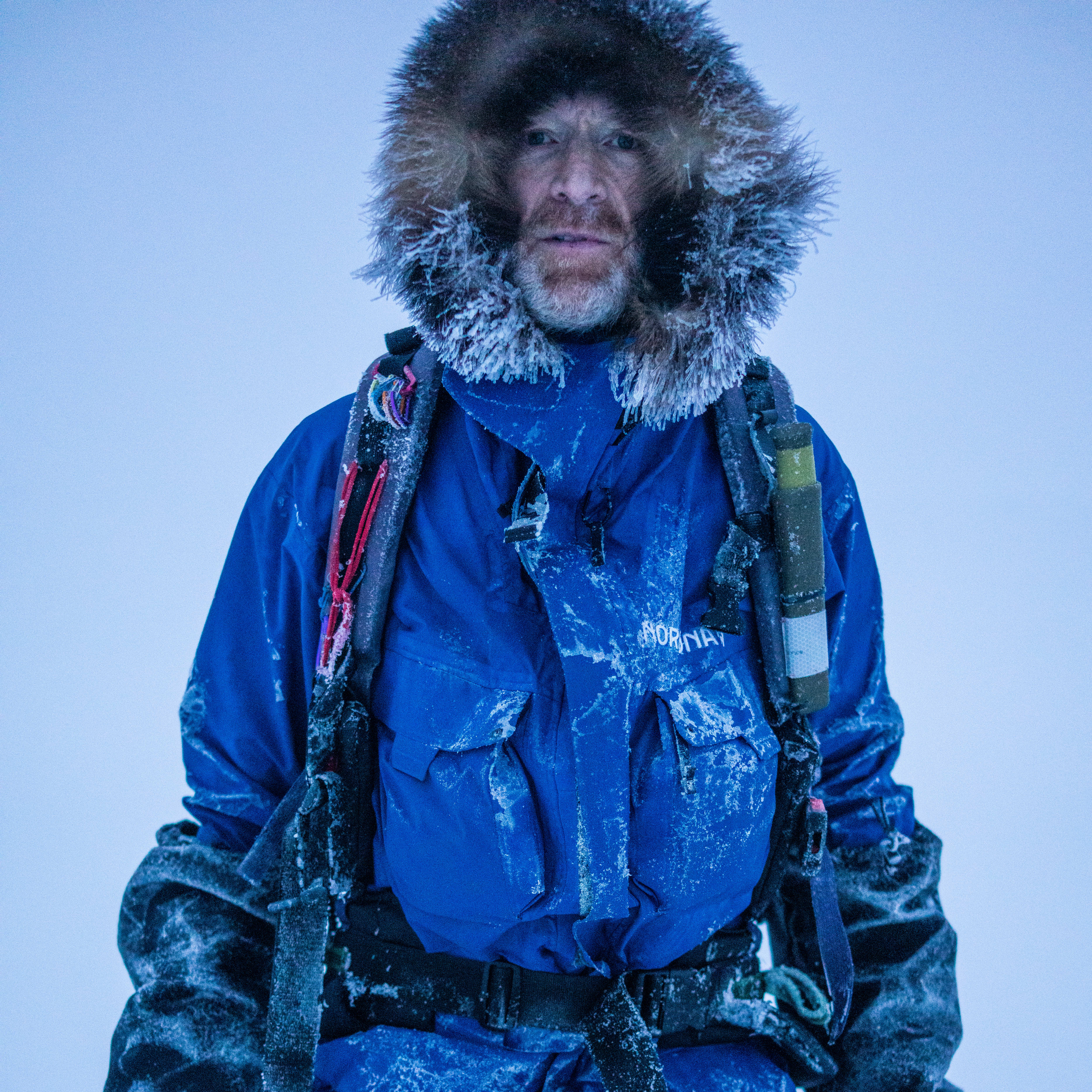 A man in a blue Norrøna jacket with a fur-lined hood is standing in the snow.  He is wearing gloves. 
