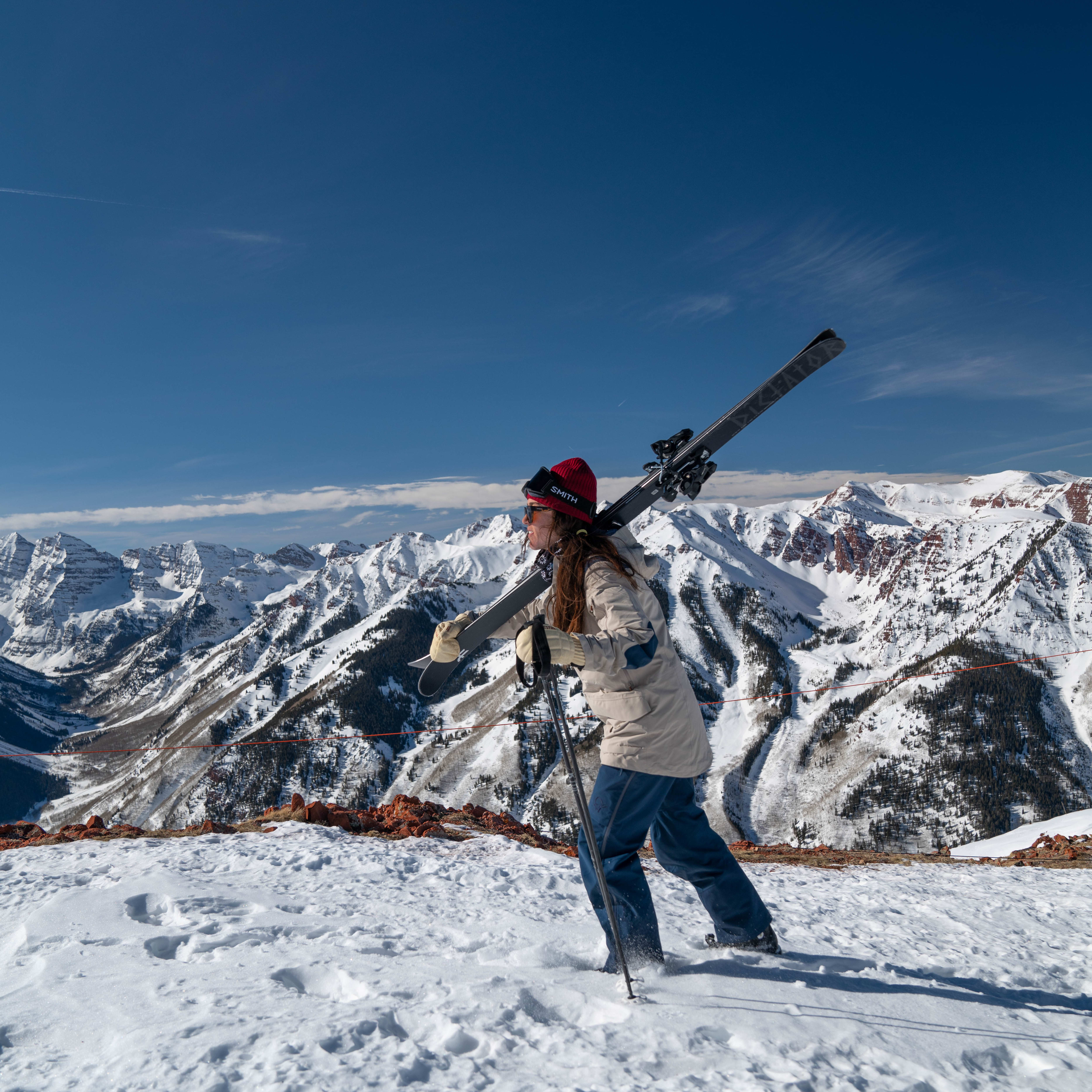 A woman in Norrøna ski pants and jacket, on a snow-covered mountain with skis on her shoulder, ski poles in hand, and snow-capped mountains in the background.