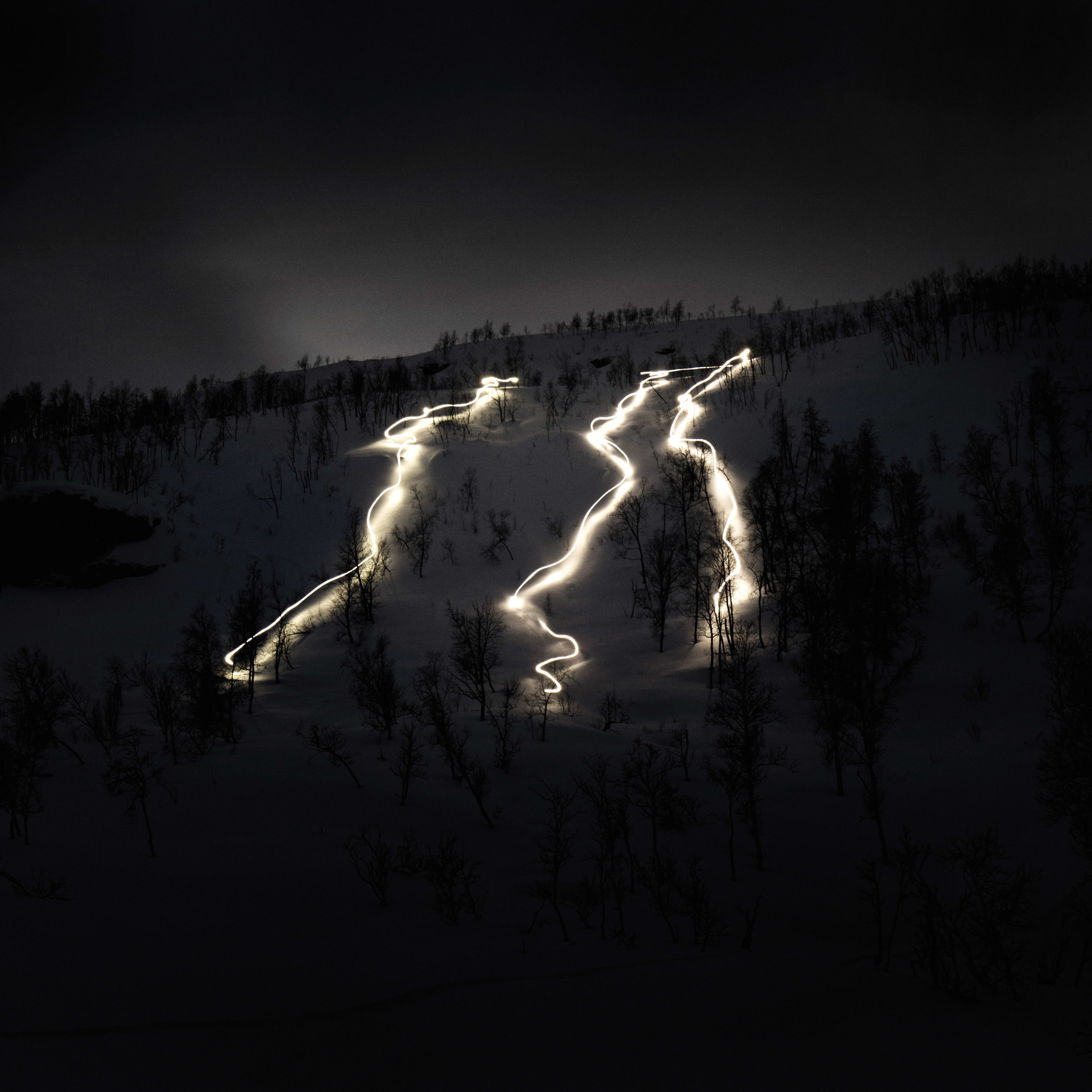 Long exposure photo of a snowy mountainside at night with three winding trails lit up by headlamps.