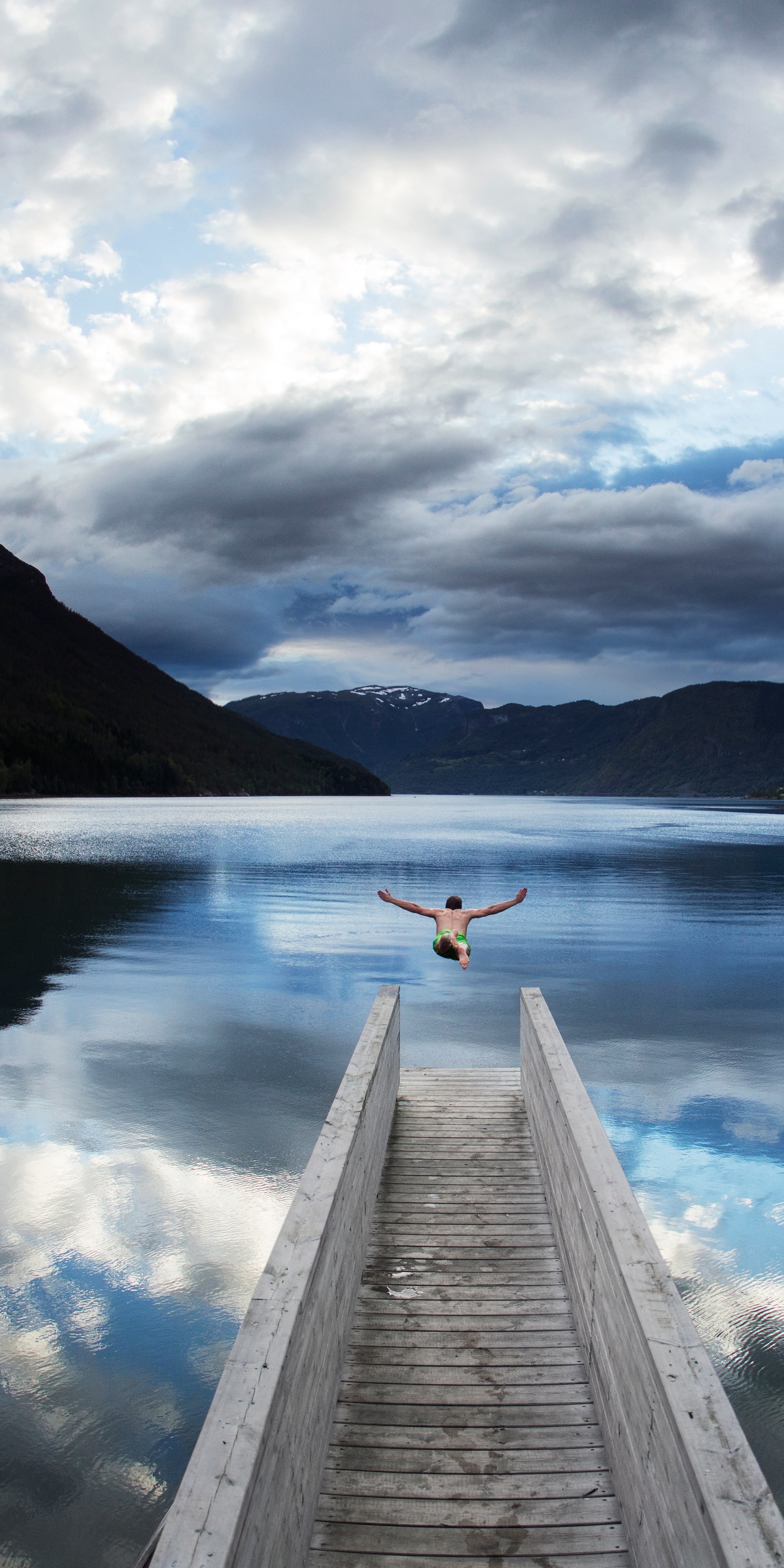 A person jumps off a wooden dock into a fjord. The sky above is partly cloudy, with the reflection of the clouds visible in the water. 
