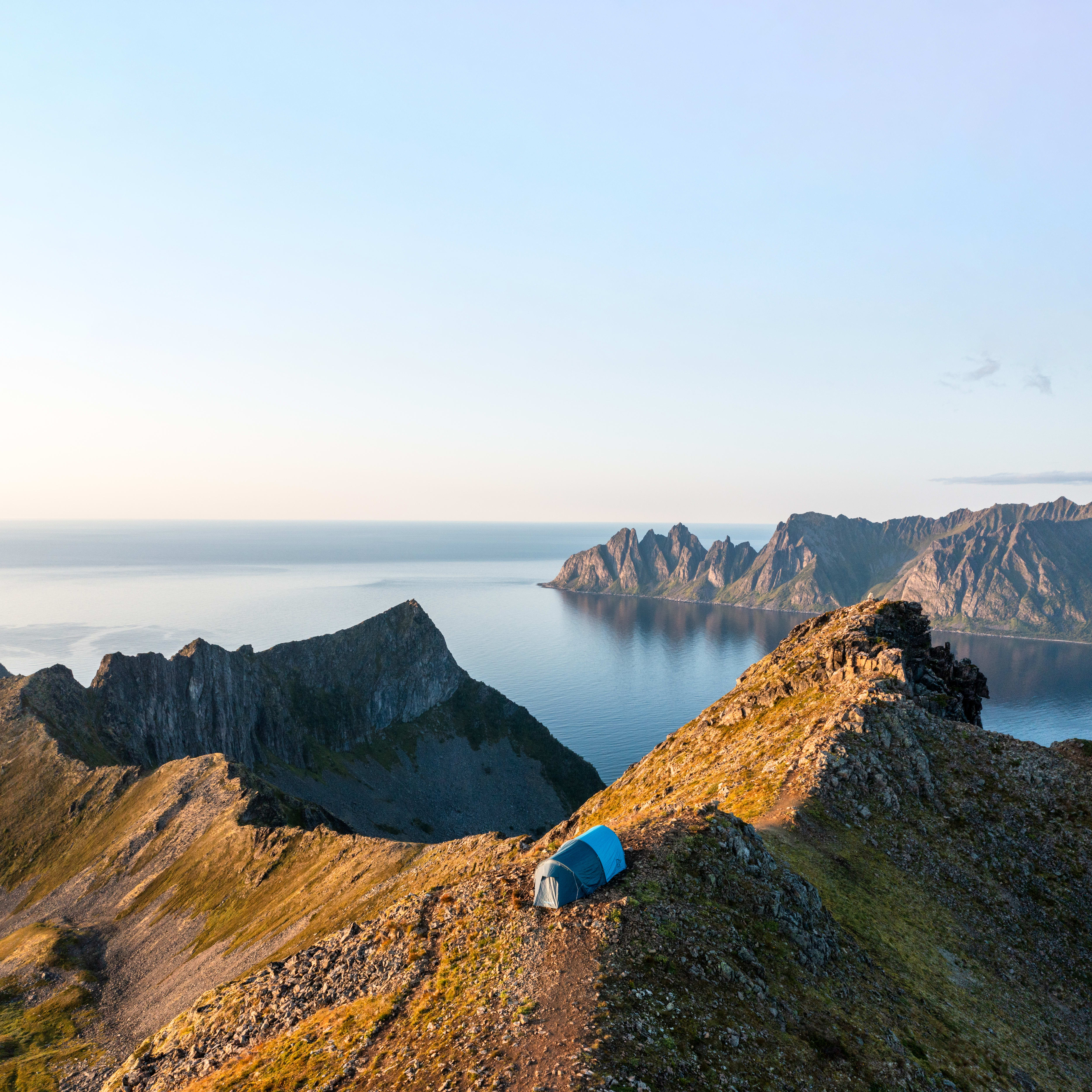 A blue Norrøna tent sits on a mountain peak overlooking a fjord.