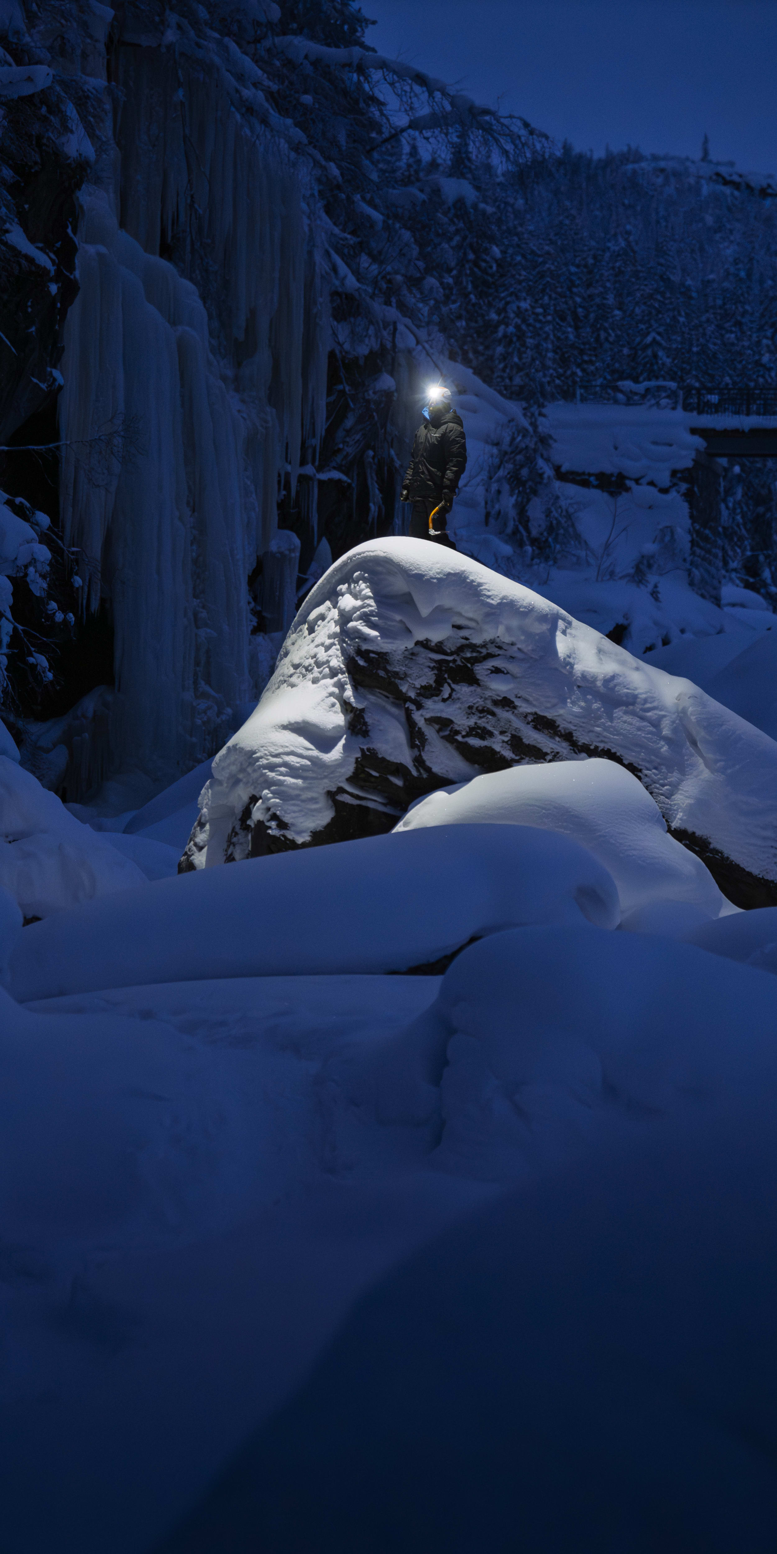 A person in a Norrøna jacket stands on a snow-covered rock wearing a headlamp at night. Ice clings to nearby cliffs.