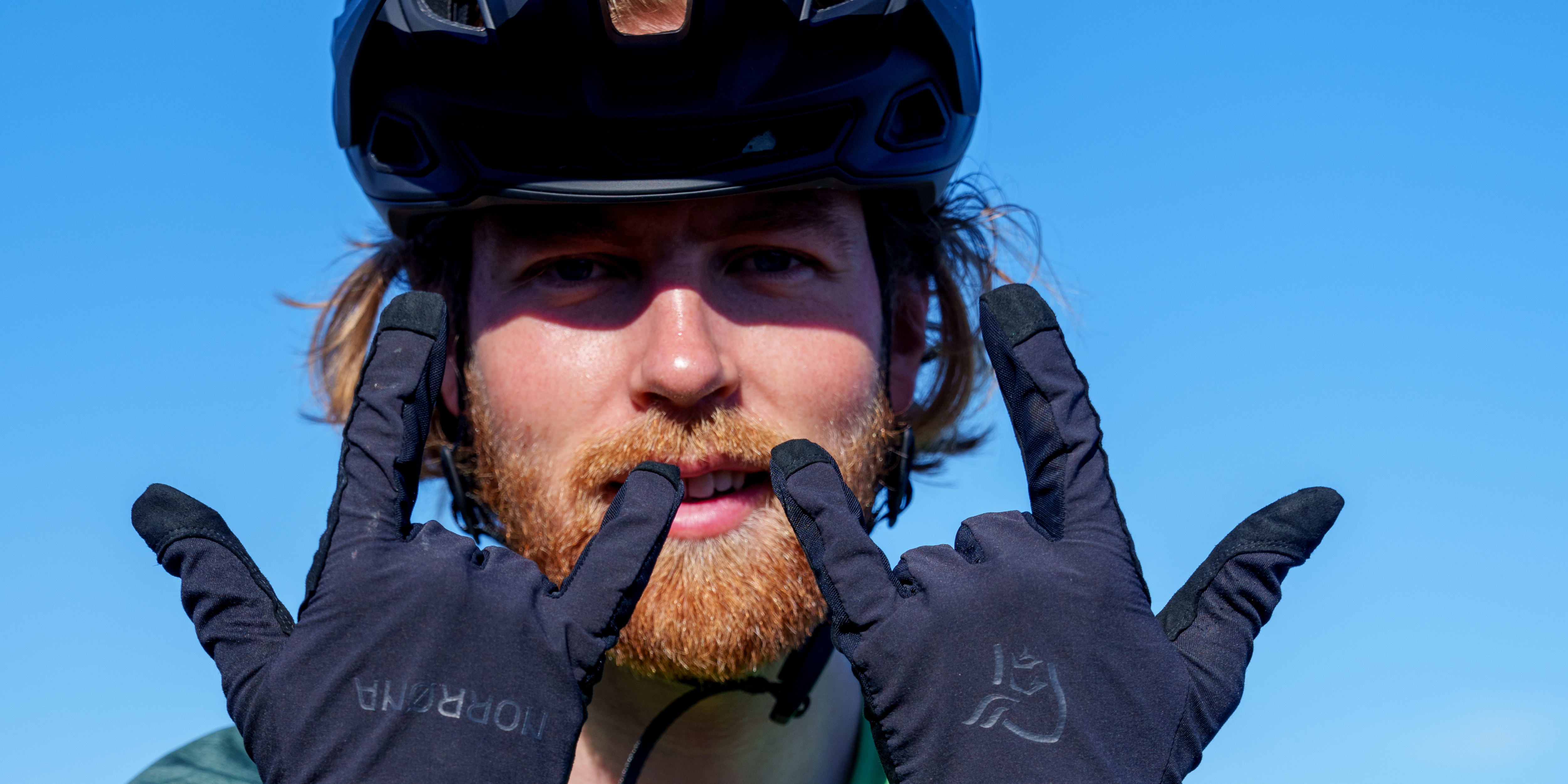 Man in a helmet and green Norrøna shirt making hand gestures with black Norrøna gloves.