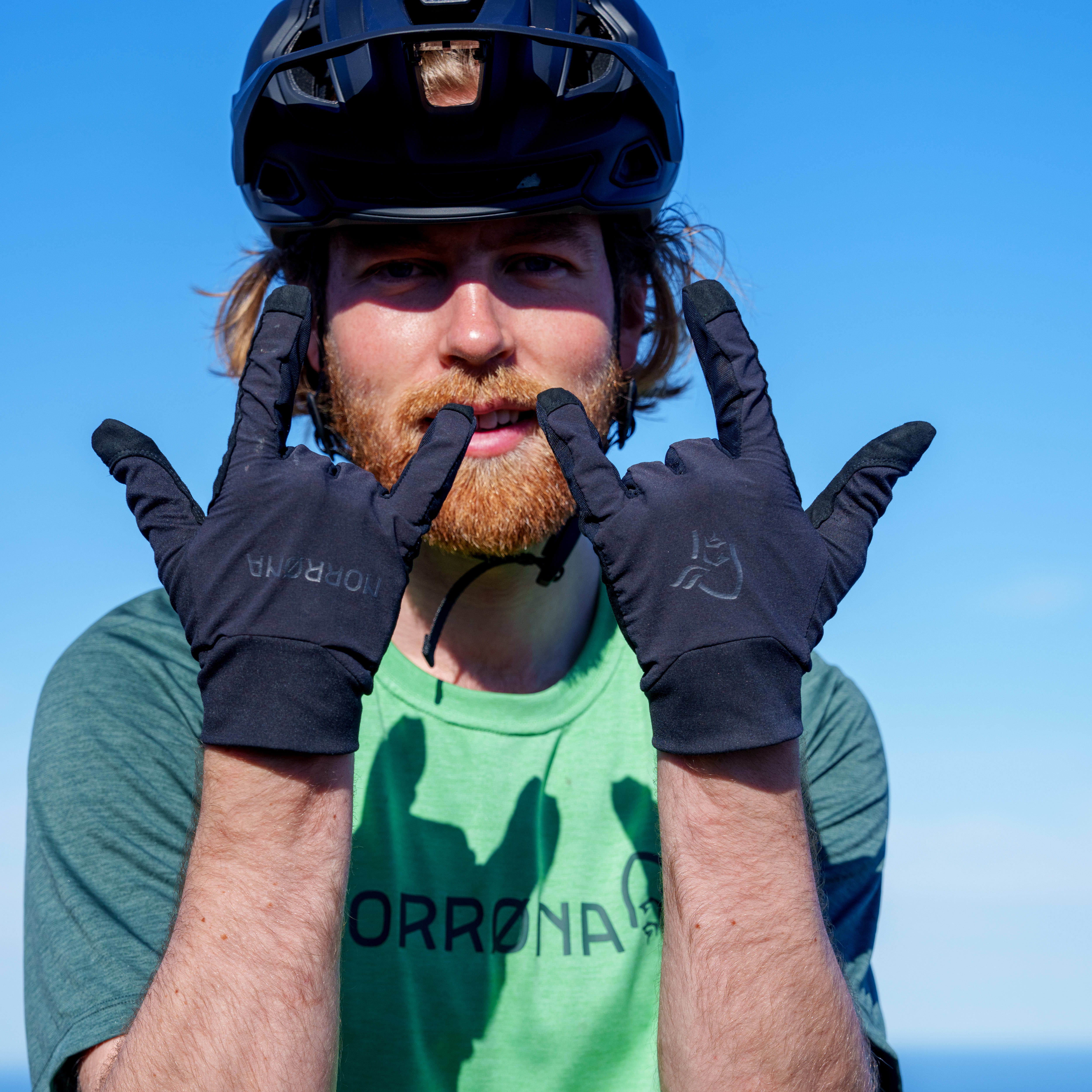 Man in a helmet and green Norrøna shirt making hand gestures with black Norrøna gloves.