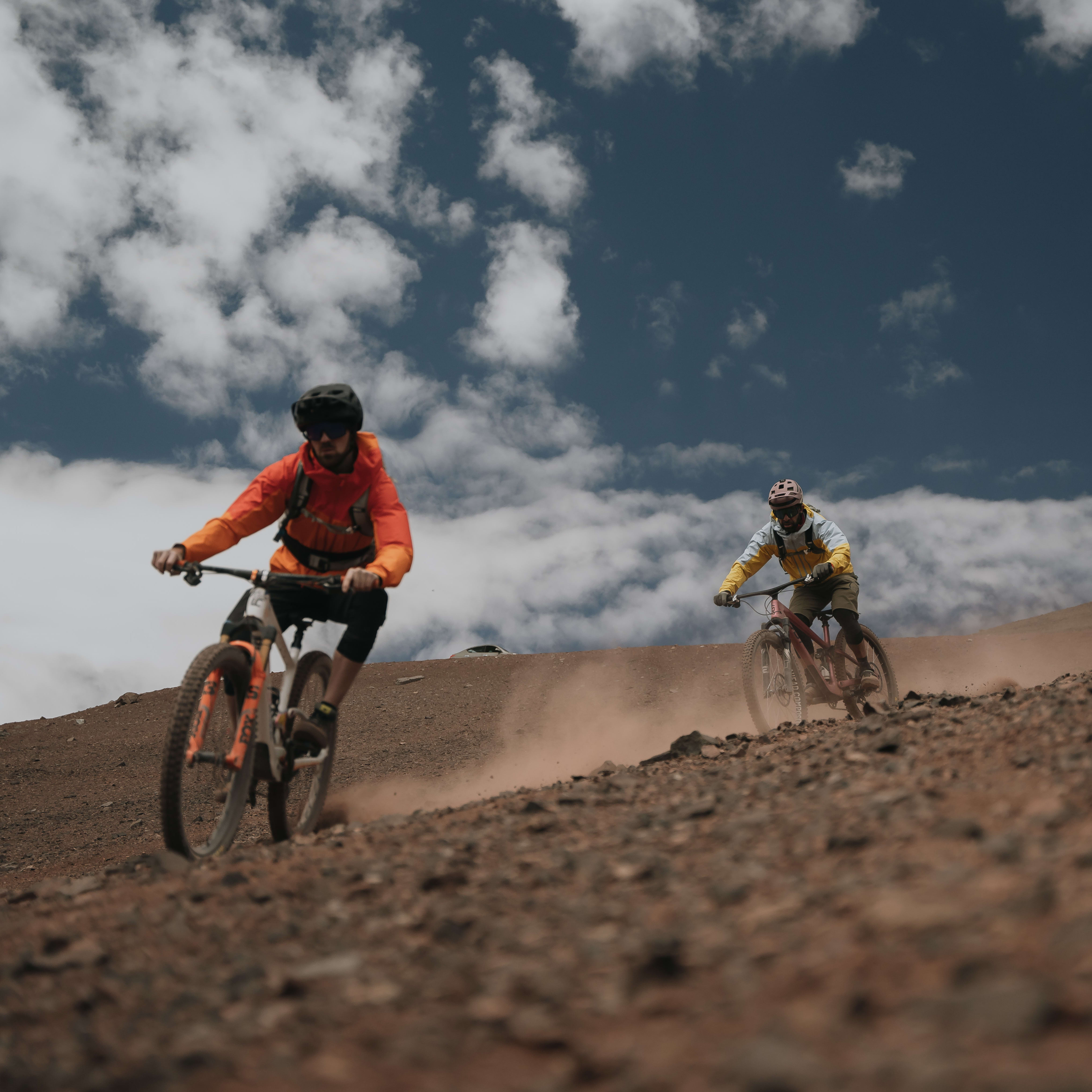 Two mountain bikers ride up a rocky trail. The biker in the foreground is wearing a Norrøna jacket.  The sky is blue with white clouds. 
