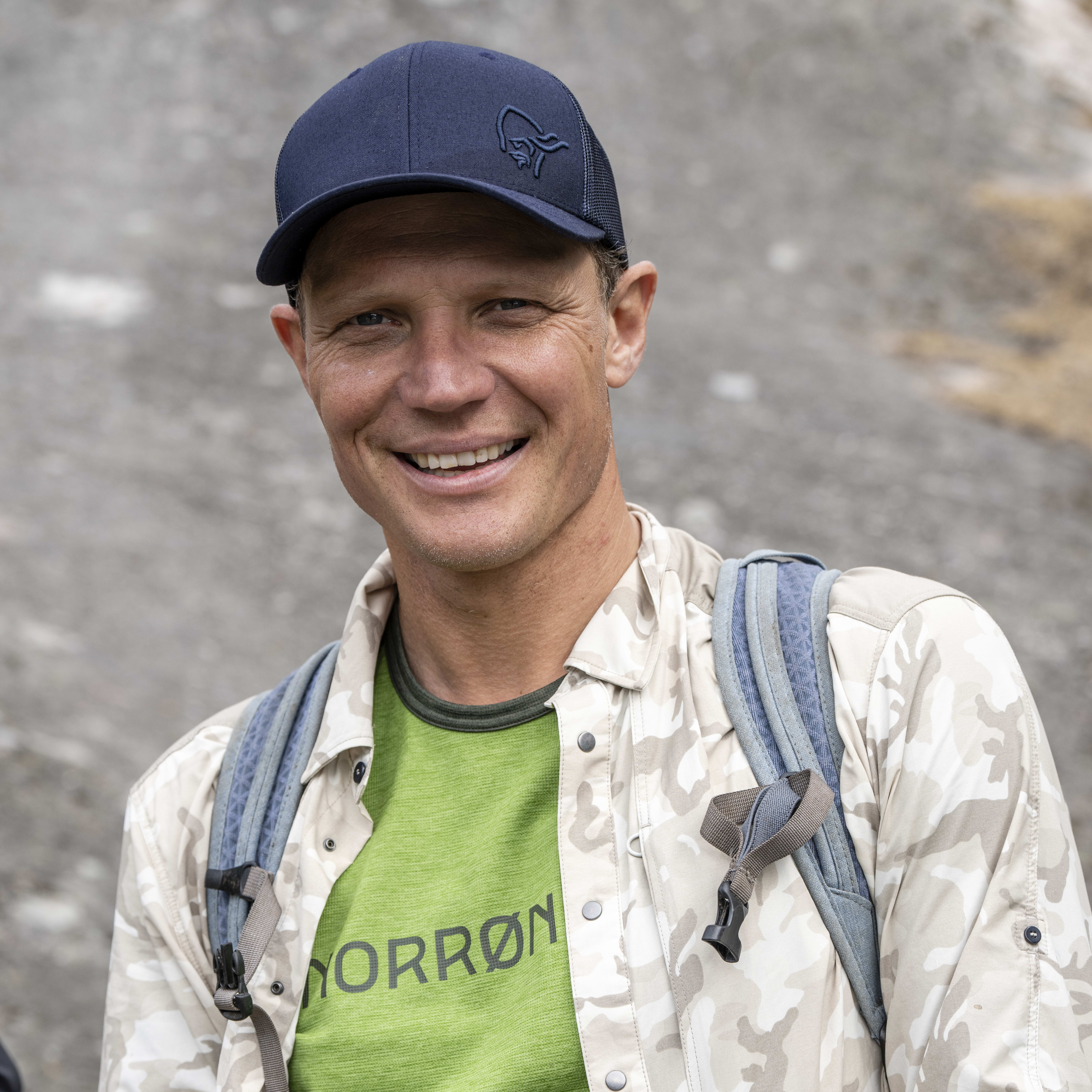 A man wearing a camouflage shirt, green t-shirt, and black gloves smiles at the camera while putting on his gloves. He is wearing a blue hat with a logo and a backpack.