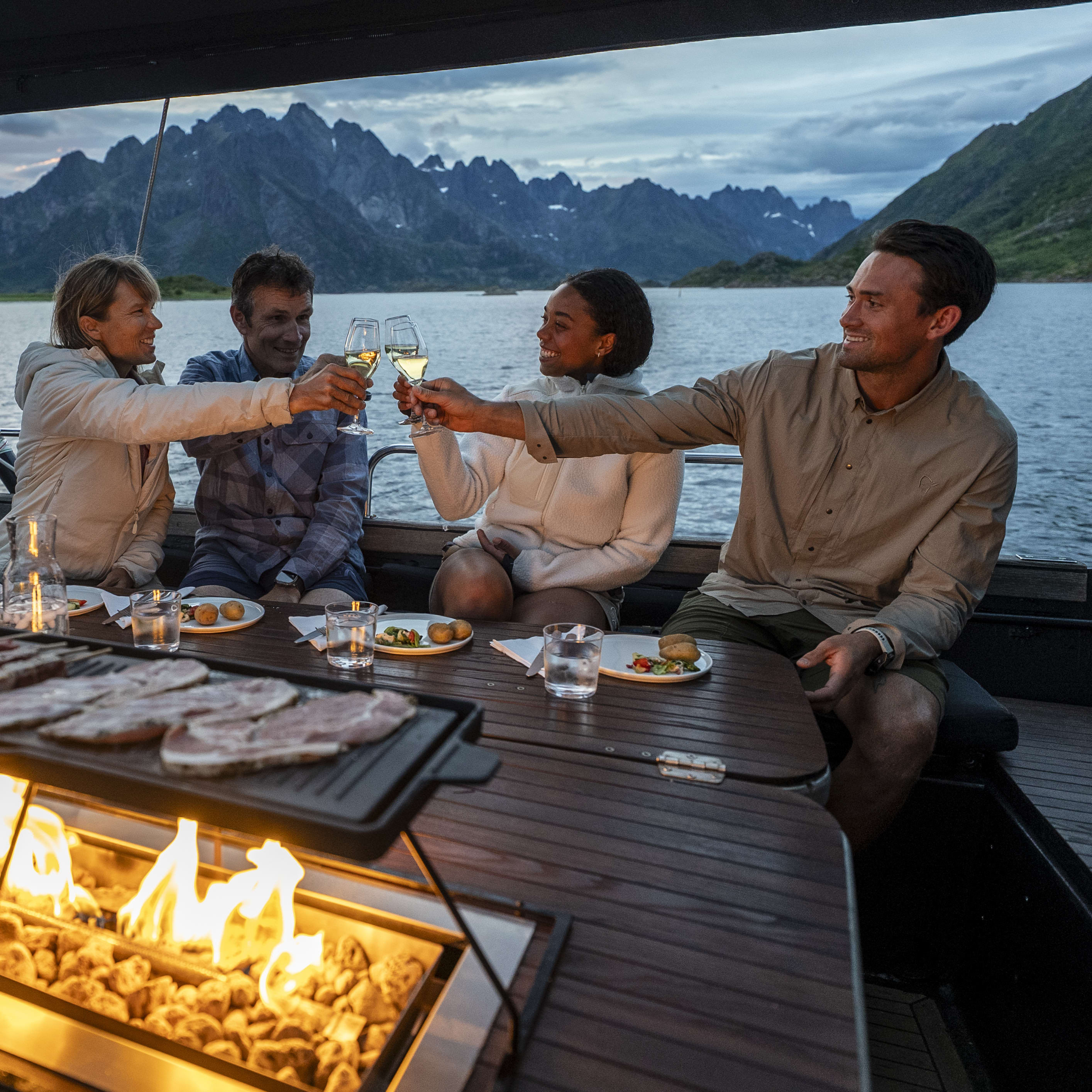 Friends toasting drinks on a boat, grilling meat, with a mountain view. The woman wears a Norrøna jacket.